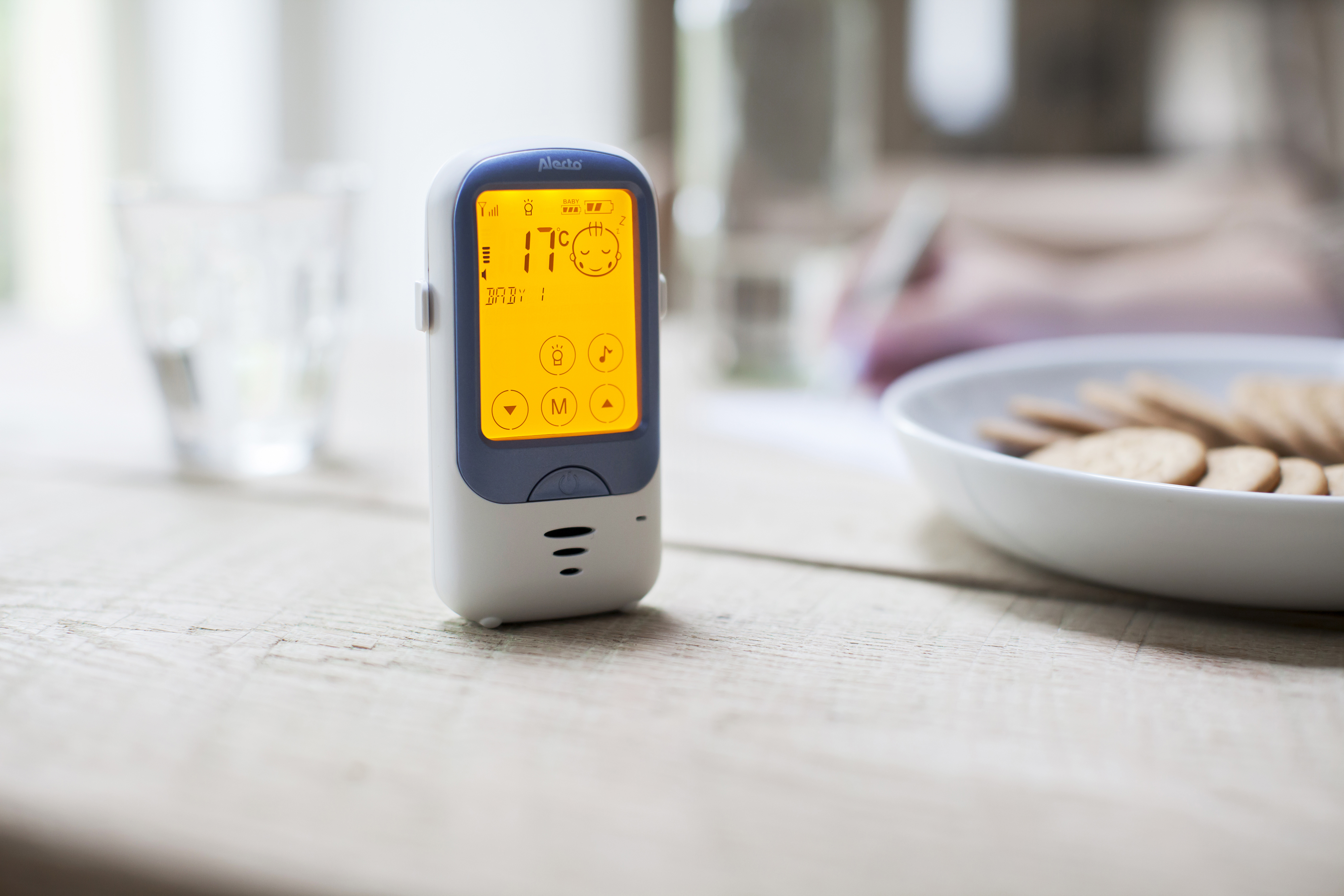Een babyfoon met een scherm dat 17°C aangeeft, staat op een houten tafel. Een glas en koekjes zijn in de buurt.