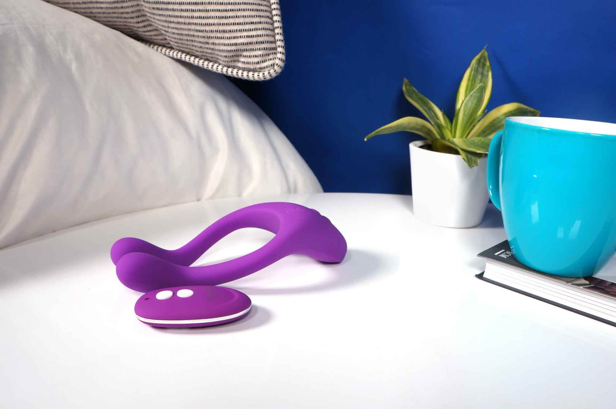 Lila Sexspielzeug und Fernbedienung auf einem weißen Tisch. Ein weißes Kissen, eine Pflanze und eine türkisfarbene Tasse sind im Hintergrund.