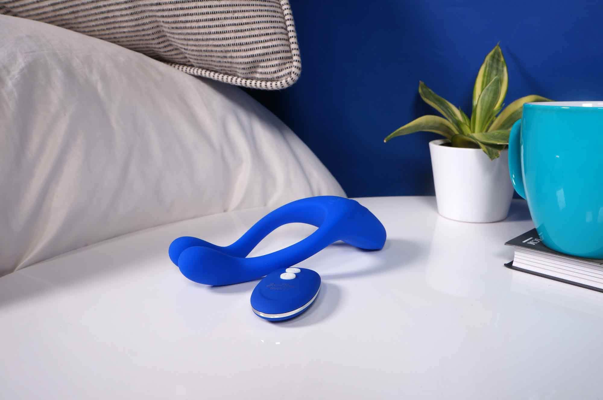 Niebieska zabawka erotyczna i pilot na białej szafce nocnej, obok poduszki, rośliny i kubka.