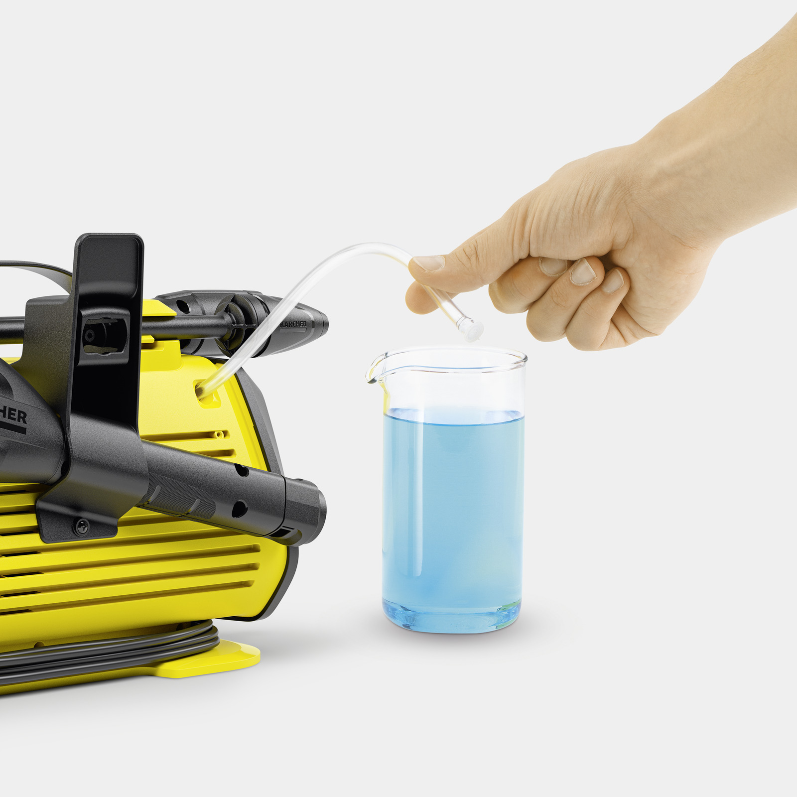 Een hand giet blauwe vloeistof in een glazen beker. Een geel-zwarte machine staat op de achtergrond. Witte achtergrond.