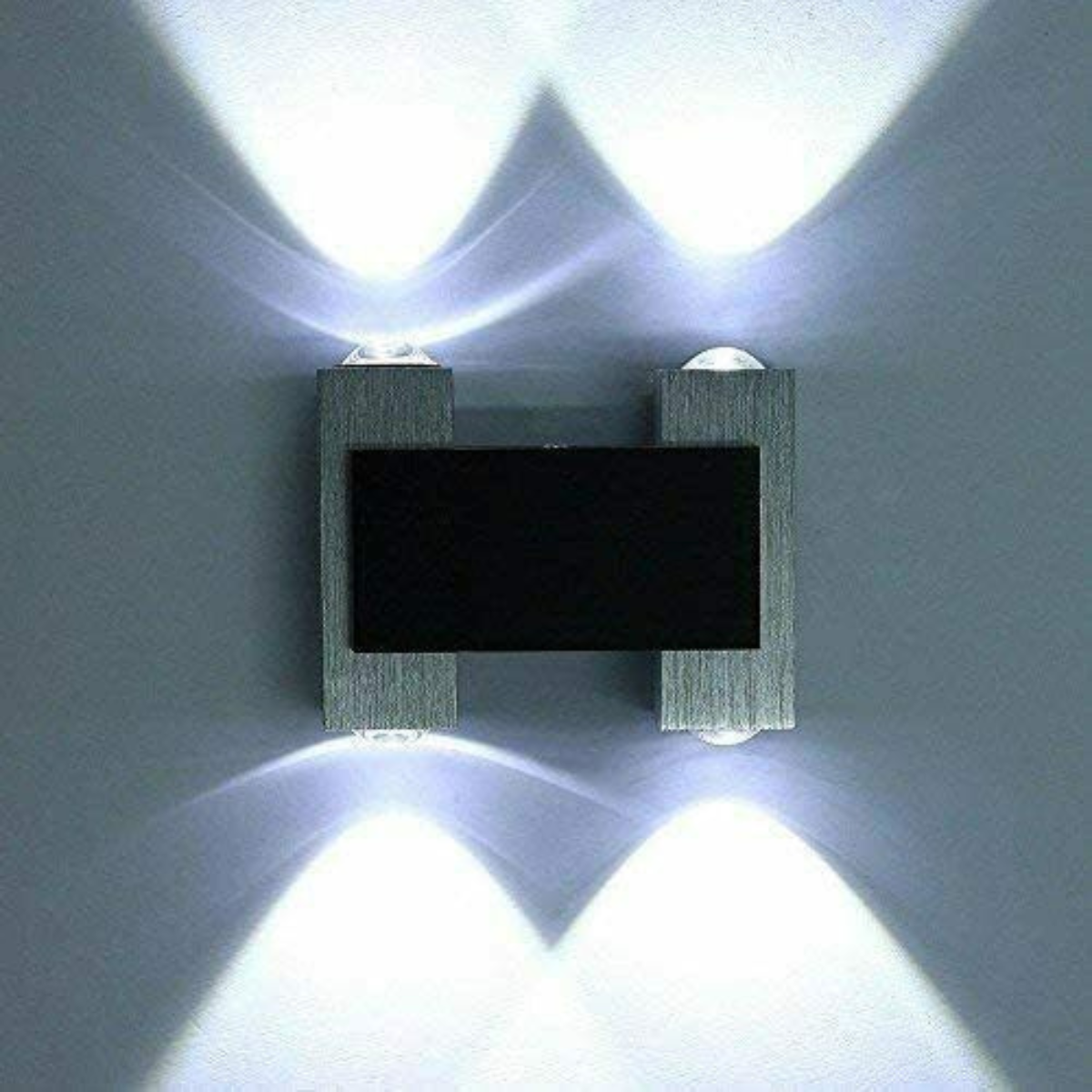 Wandleuchte mit rechteckigem schwarzem Mittelteil, silbernen Seiten und hellen Lichtstrahlen an einer grauen Wand.