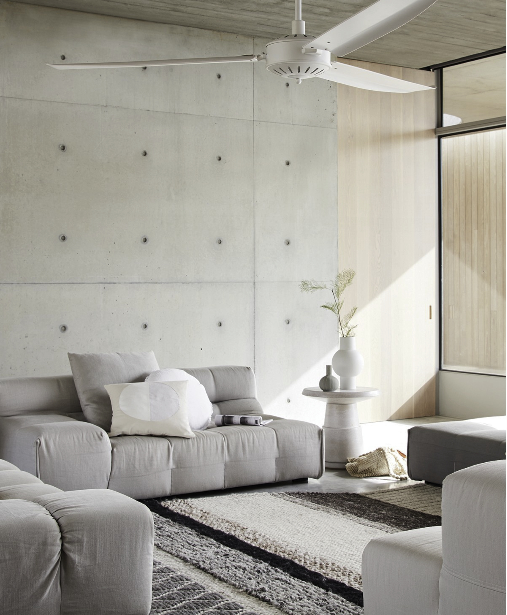 Ein modernes Wohnzimmer mit hellgrauem Sofa, Teppich und Deckenventilator. Betonwand-Hintergrund.