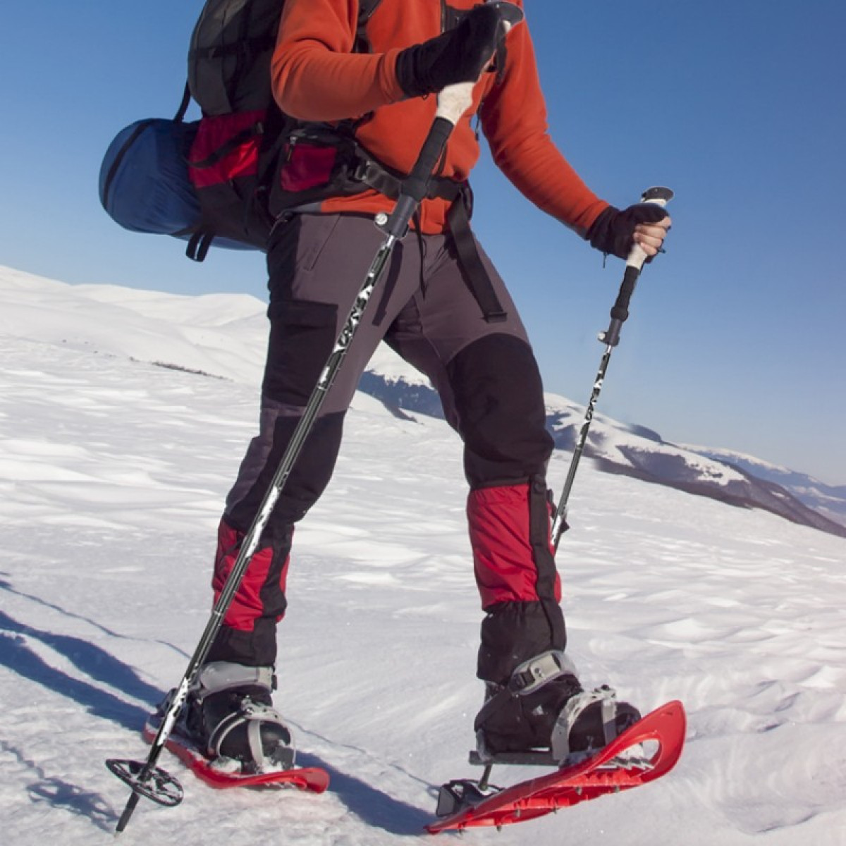 Osoba w rakietach śnieżnych idąca po śniegu. Kije i plecak widoczne na tle niebieskiego nieba.