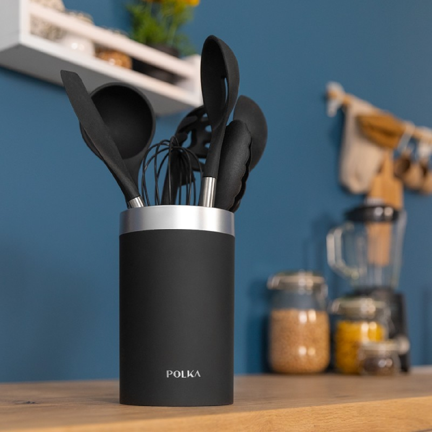 Utensili da cucina in un supporto nero su un piano di lavoro in legno, con parete blu e barattoli.