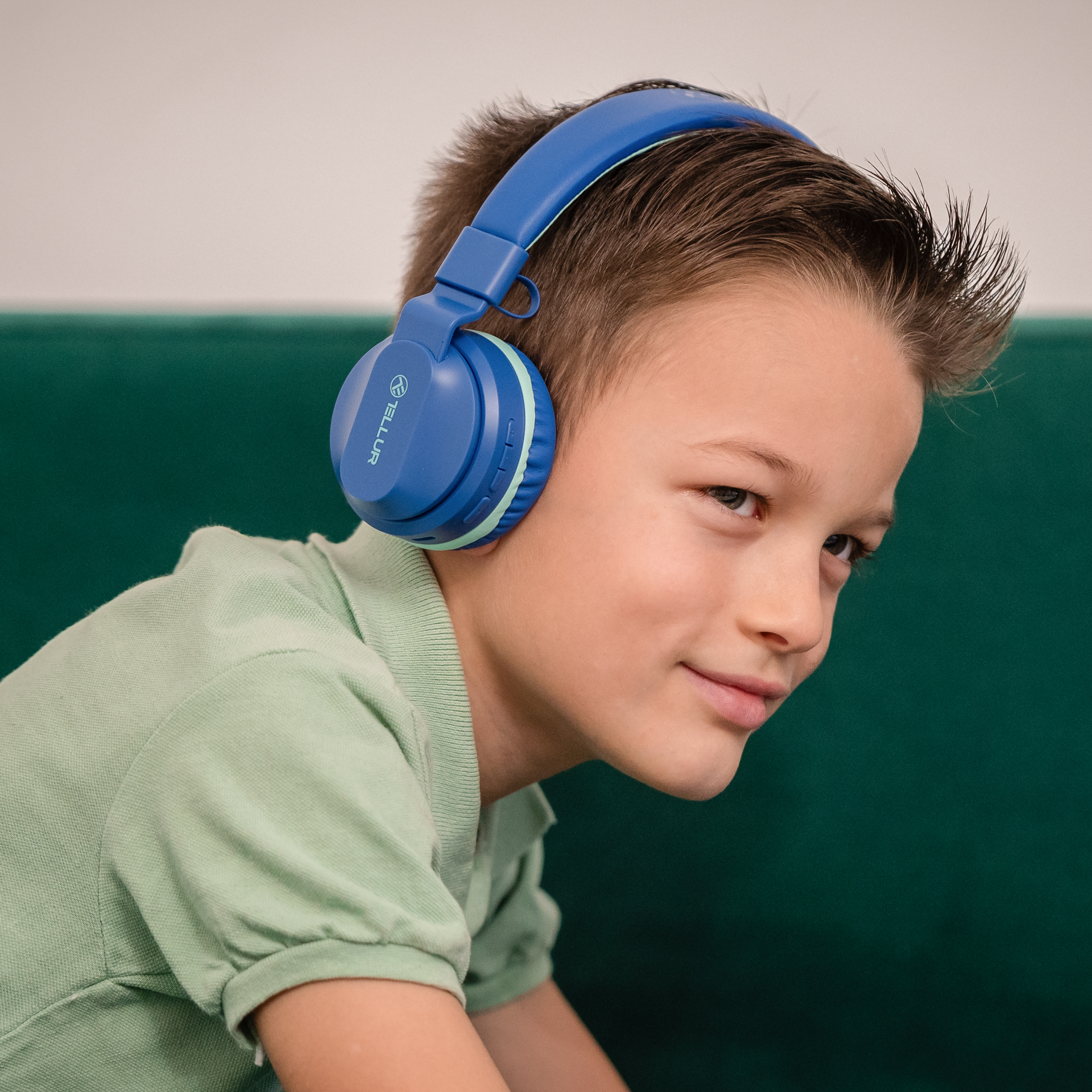 Młody chłopiec w niebieskich słuchawkach, patrzący w prawo, uśmiechnięty. Zielone tło.