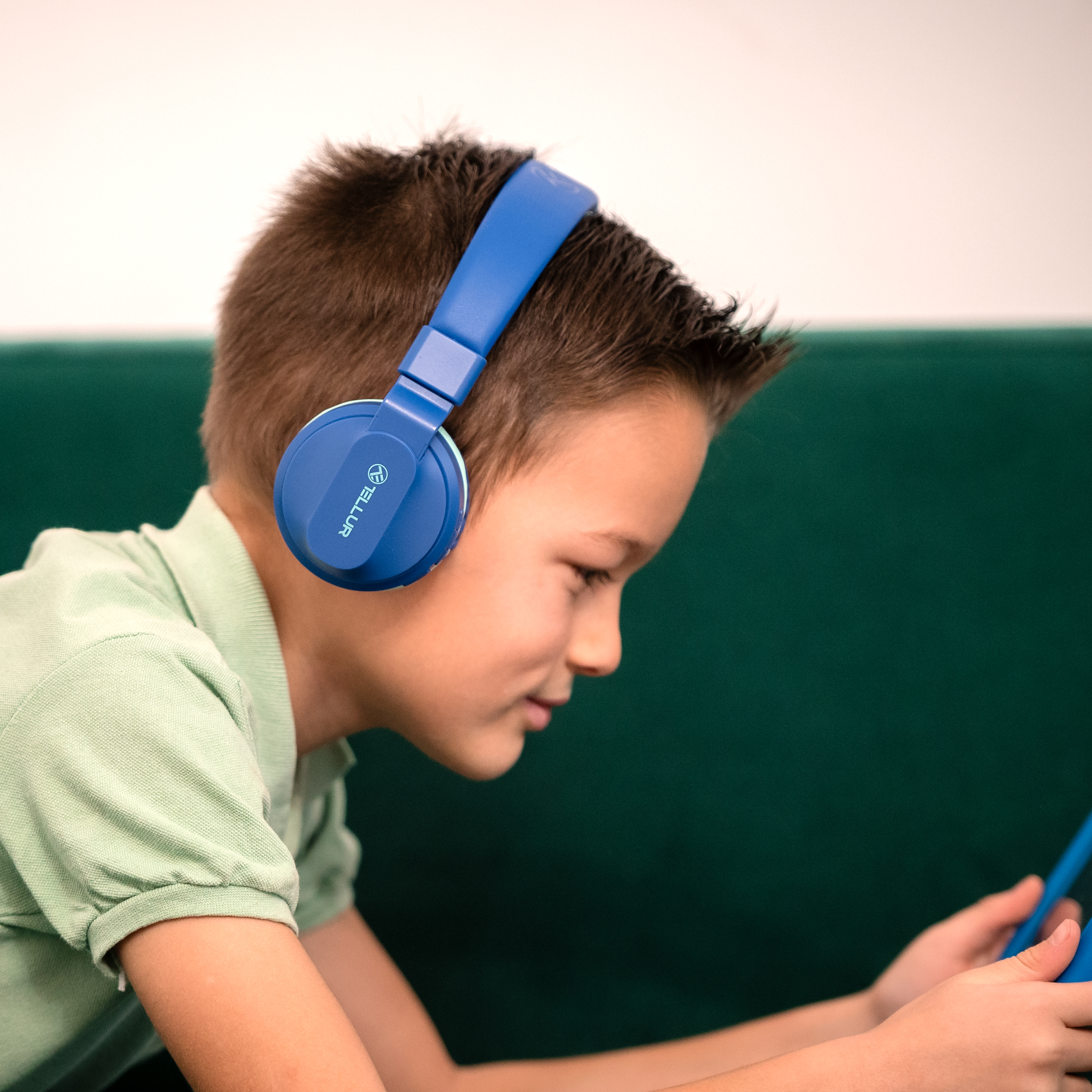 Młody chłopiec z niebieskimi słuchawkami skupia się na tablecie. Ma na sobie zieloną koszulę.