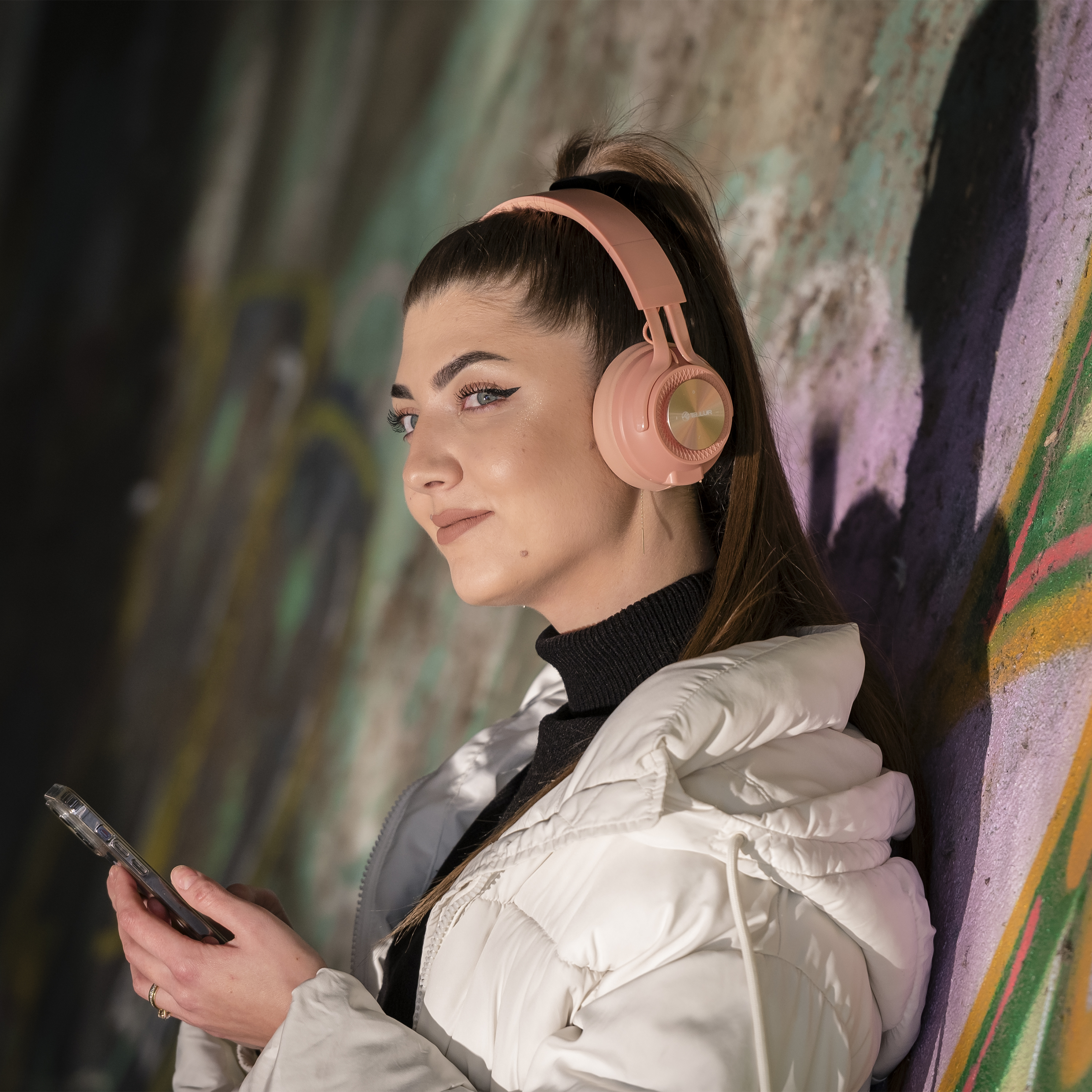Frau mit rosa Kopfhörern, hält ein Telefon, lehnt an einer Graffiti-Wand.