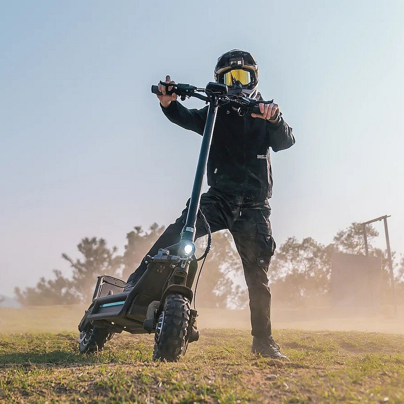 Person mit Helm und Brille auf einem Roller auf einem Feld, das Staub aufwirbelt. Sonniger Tag.