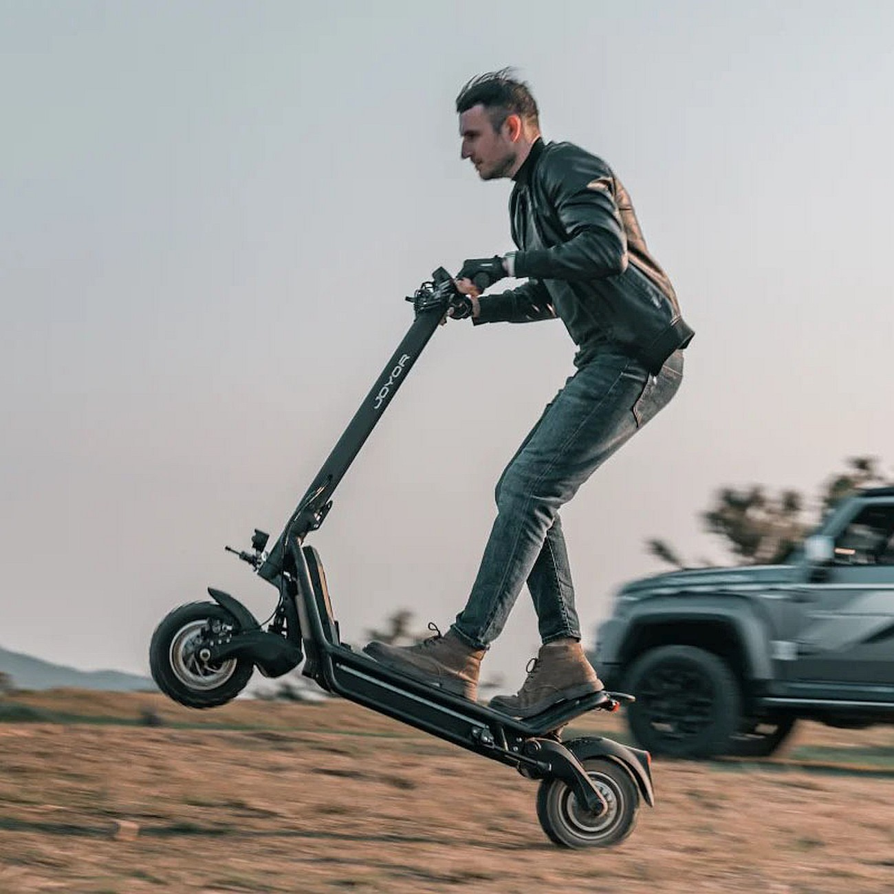 Mann fährt Joyor-Roller, Vorderrad oben, auf Feldweg, Auto im Hintergrund.