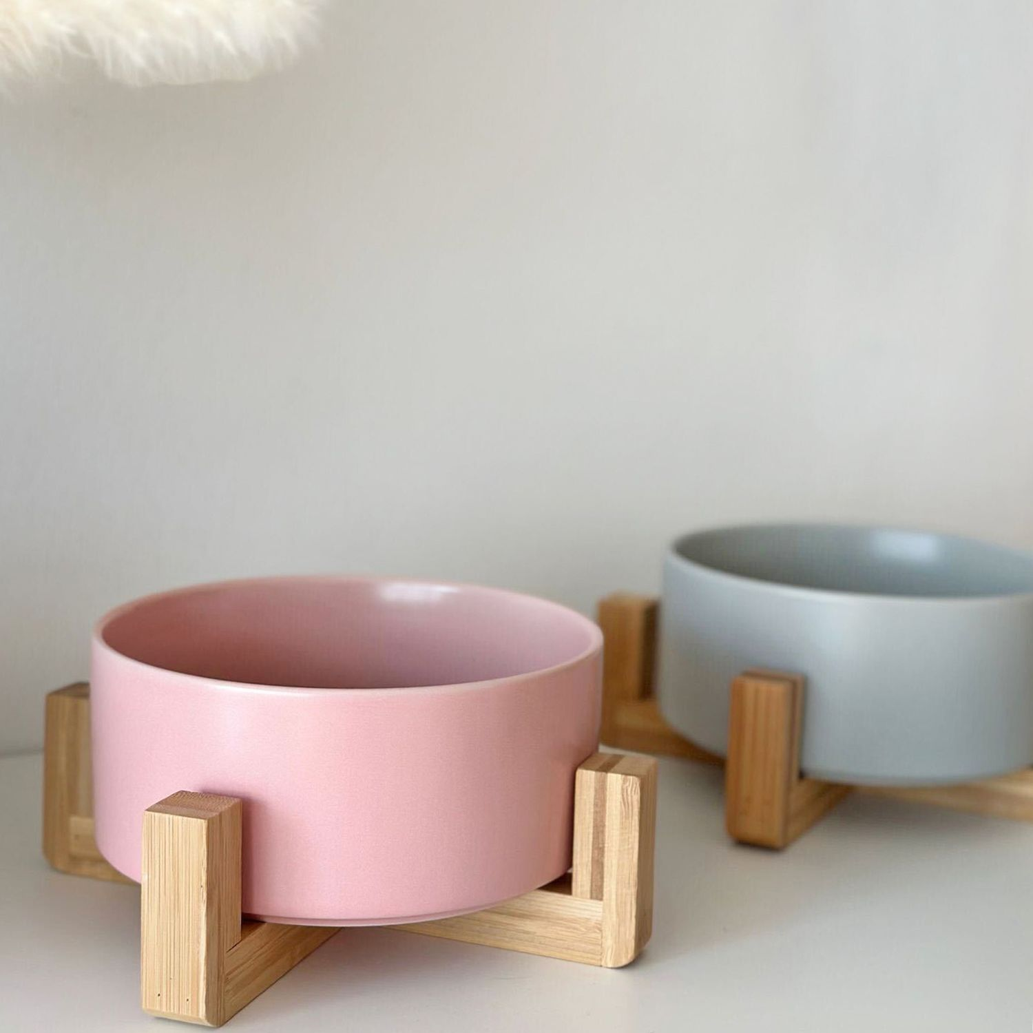 Zwei Keramik-Futternäpfe, rosa und grau, auf Holzständern. Weiße Wand im Hintergrund, mit einem flauschigen Objekt.