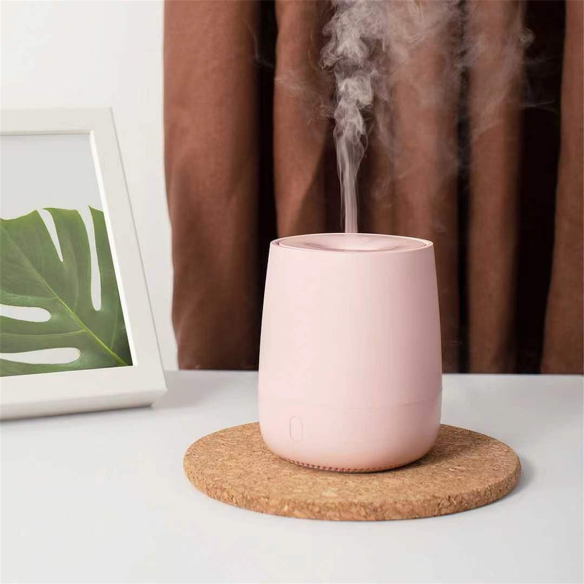 Ein rosa Luftbefeuchter mit Dampf, auf Korkunterlage, neben einem Bild auf weißem Tisch.