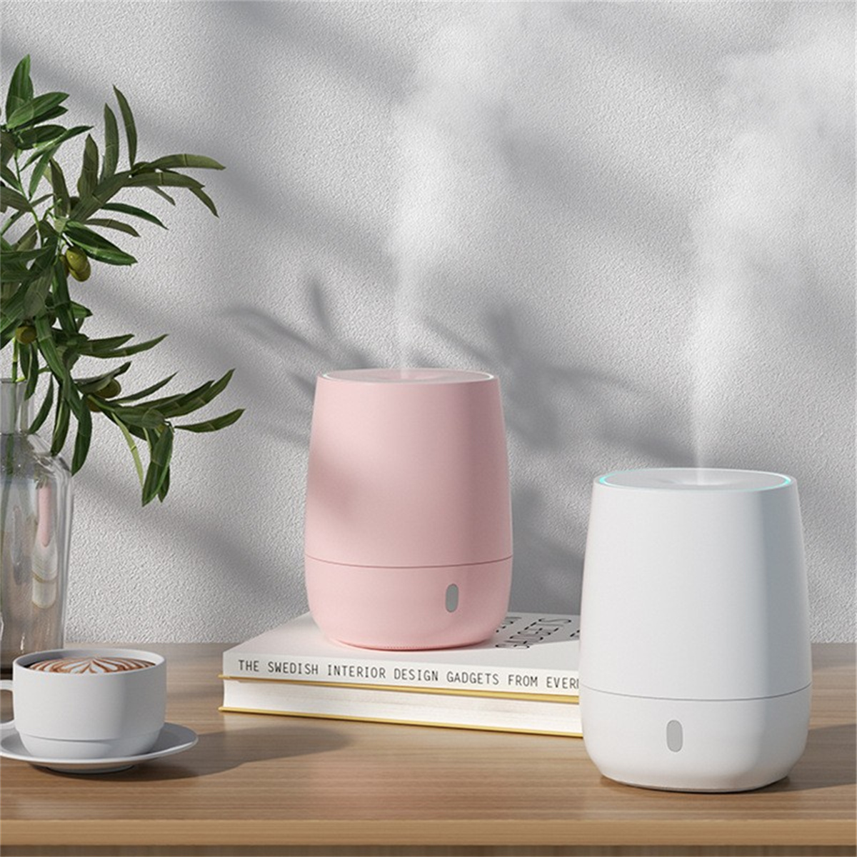 Zwei zylindrische Diffusoren, rosa und weiß, auf einem Holztisch mit Kaffeetasse und Pflanze.