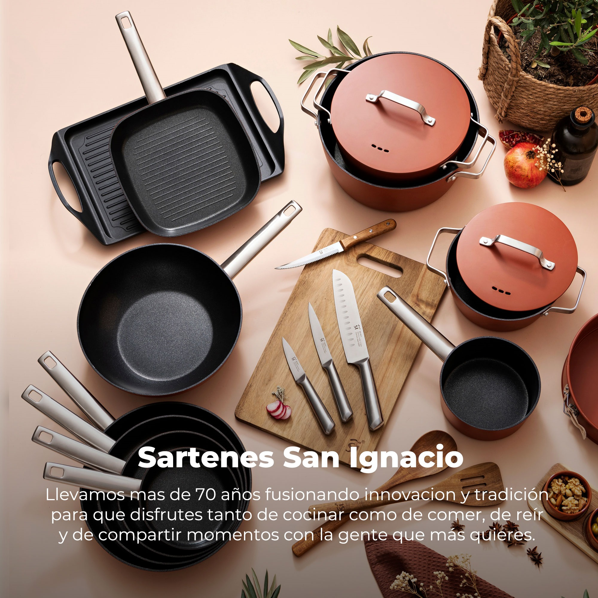 Juego de utensilios de cocina con sartenes, ollas y tapas. Cuchillos y una tabla de cortar en el centro. Granada y plantas al fondo.