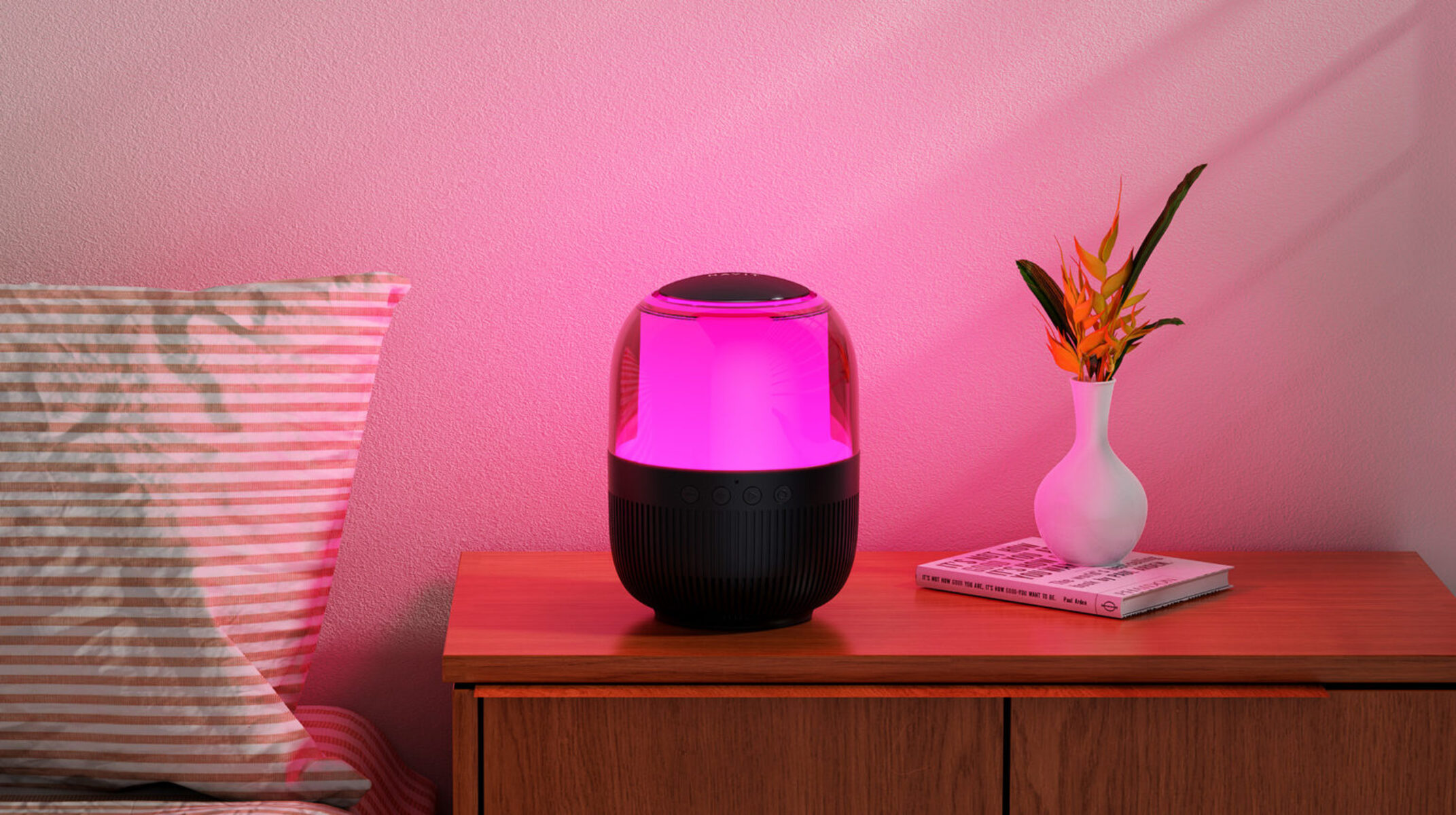 Głośnik na drewnianym stole z lampą i książką, na tle różowej ściany, tworząc przytulną scenę.