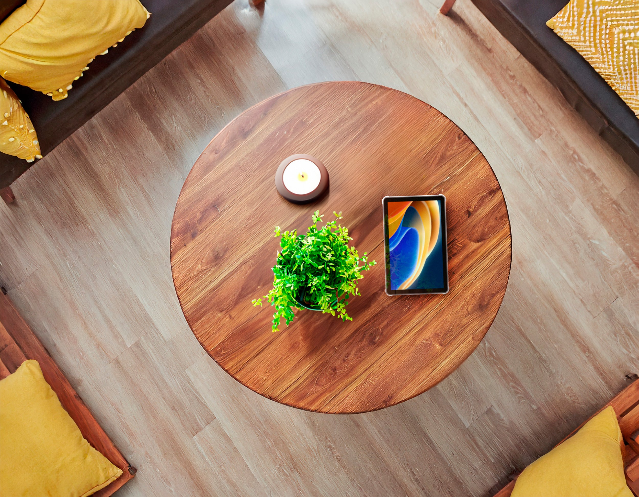 Vista superior de una mesa de madera con una vela, una planta y una tableta que muestra una pantalla colorida. Almohadas amarillas alrededor.