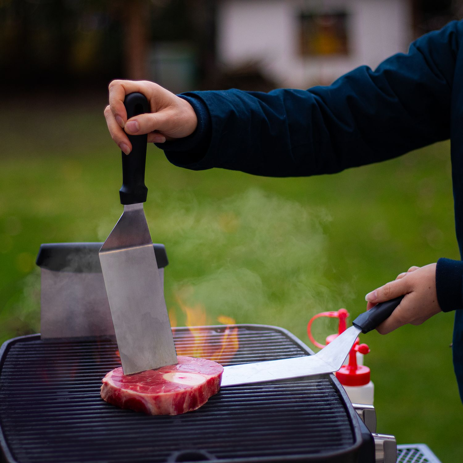 Person grillt ein Steak auf einem Grill. Rauch steigt auf. Eine Ketchupflasche und ein Pfannenwender sind sichtbar.