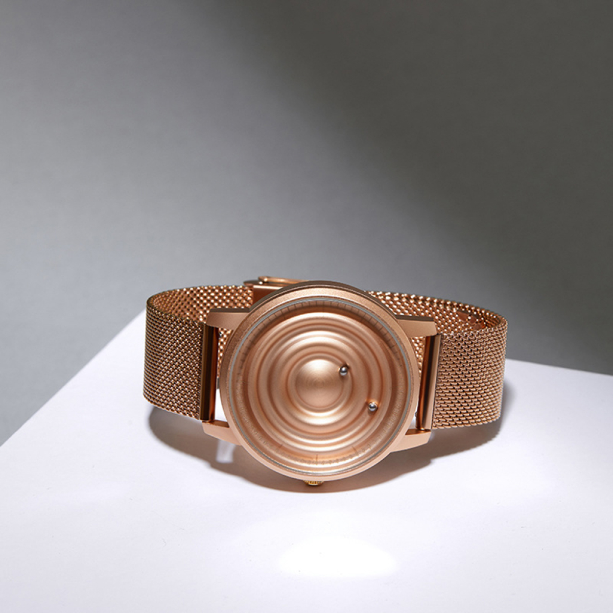 Roségoldene Uhr mit Mesh-Armband. Das Zifferblatt hat konzentrische Kreise. Auf weißer Oberfläche.