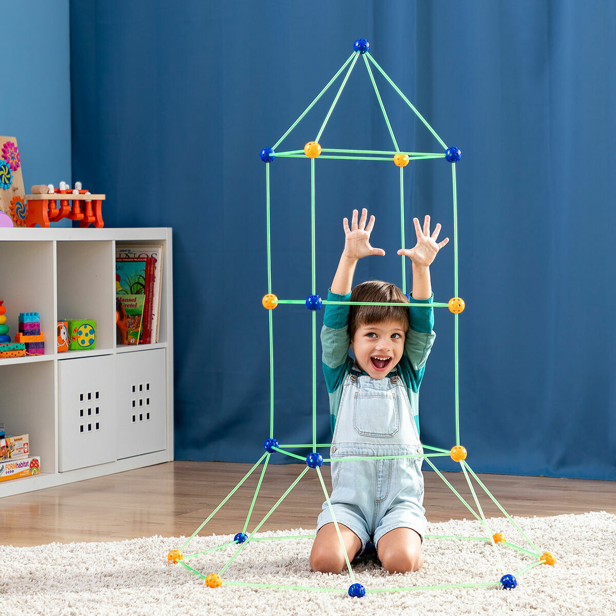 Un bambino costruisce una struttura con bastoncini verdi e connettori blu e arancioni, sorridendo alla fotocamera.