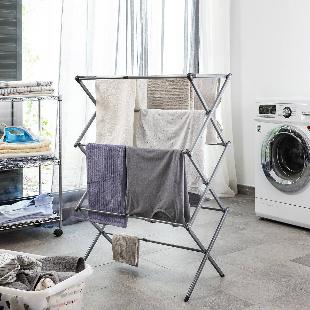 Un tendedero con ropa colgada. Al lado hay una lavadora y una cesta de ropa. La habitación está alicatada.