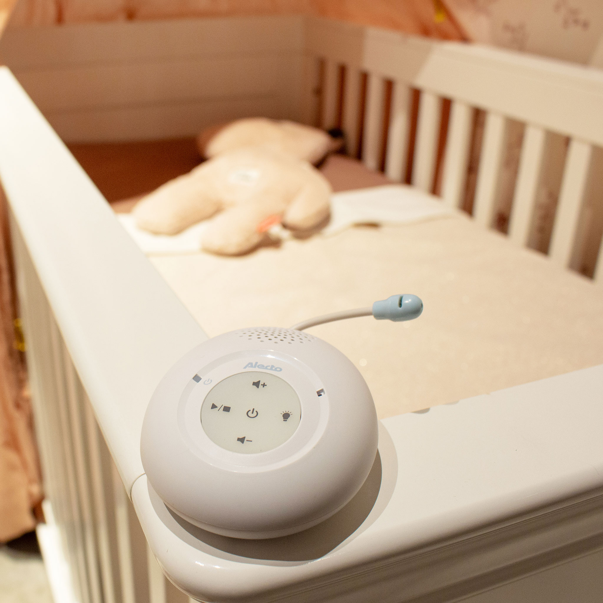 White baby monitor on a crib. A teddy bear is visible inside the crib. Pink blanket is on.