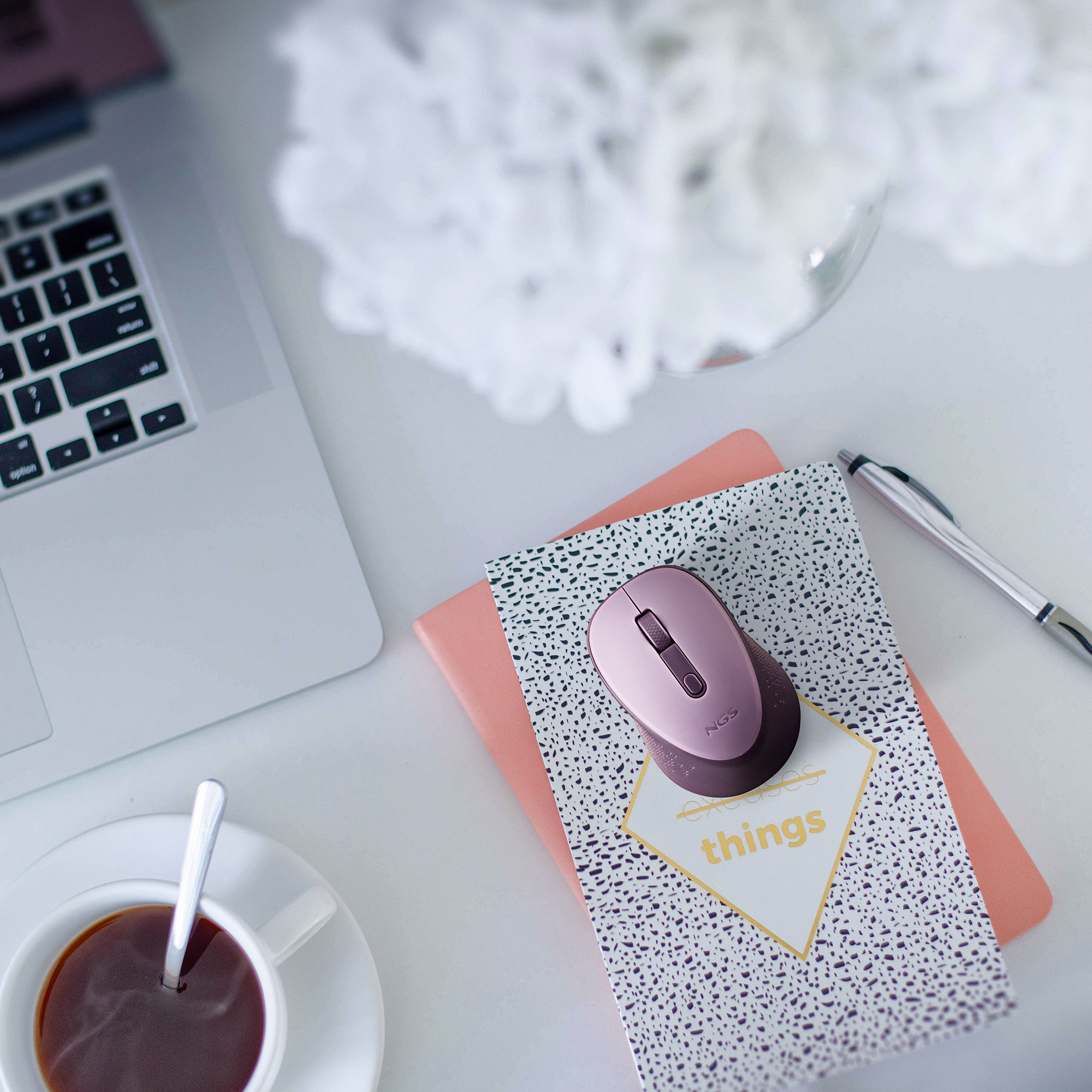 Un'impostazione della scrivania con un laptop, un mouse rosa, un blocco note, una penna e una tazza di caffè con fiori sullo sfondo.