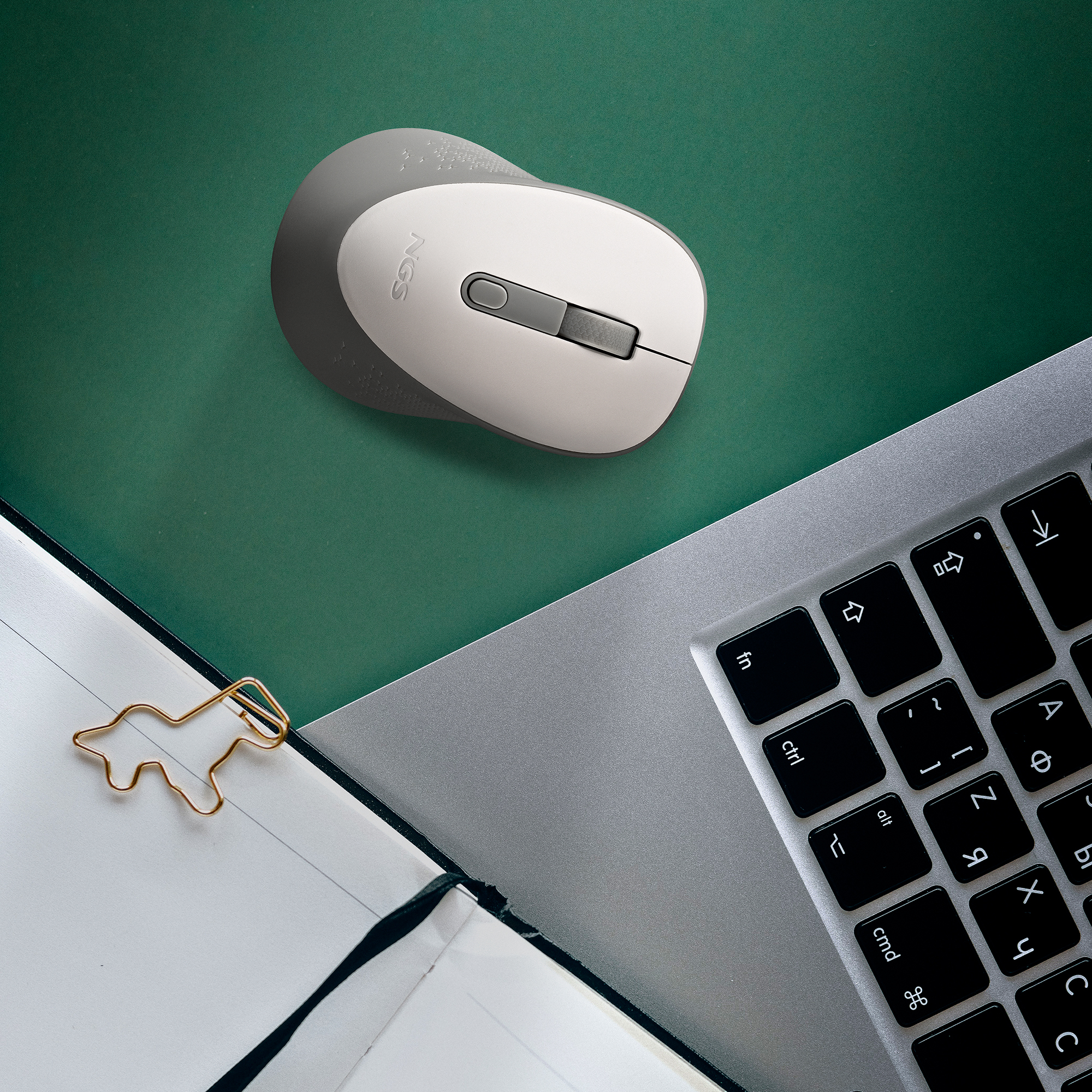 Ratón de computadora gris y blanco sobre una superficie verde, cerca de una laptop y un clip dorado.