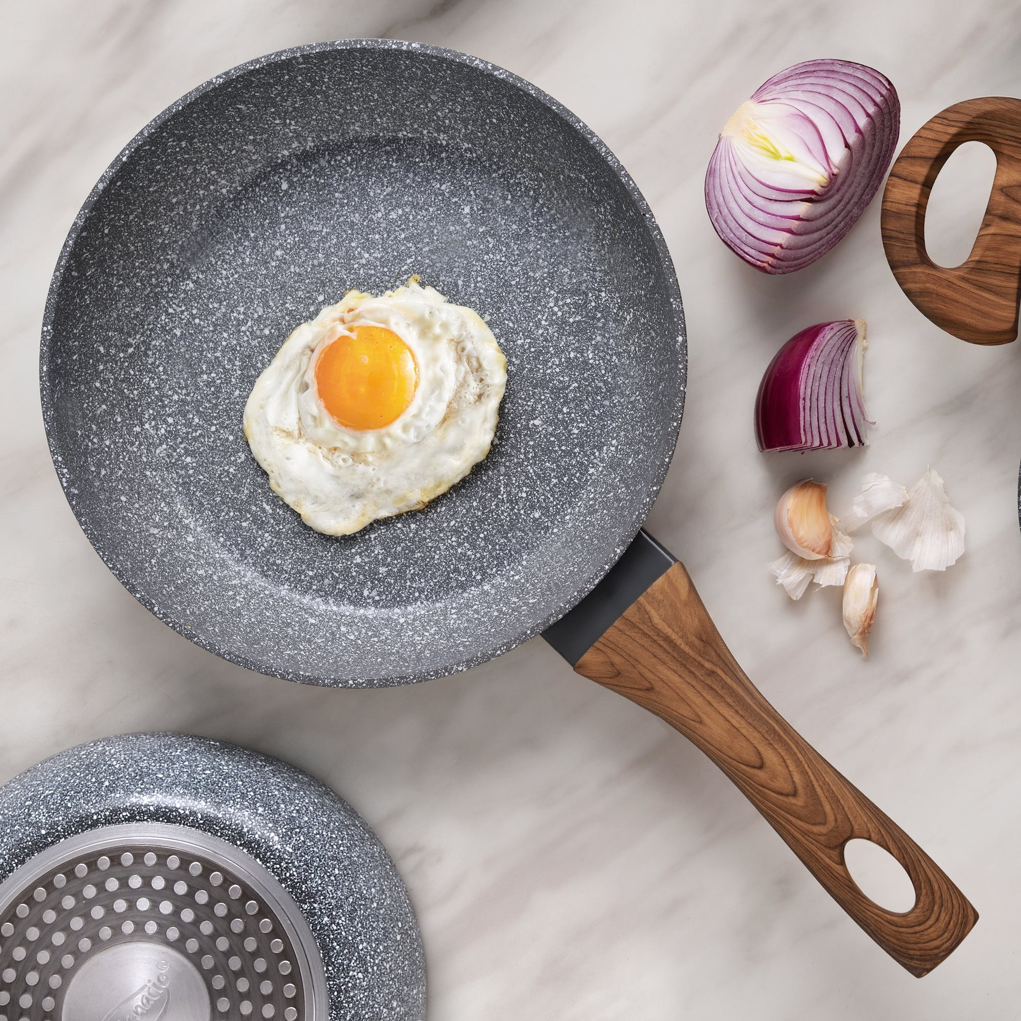 Sartén con huevo frito, cebolla roja, ajo y mango de madera sobre superficie de mármol.