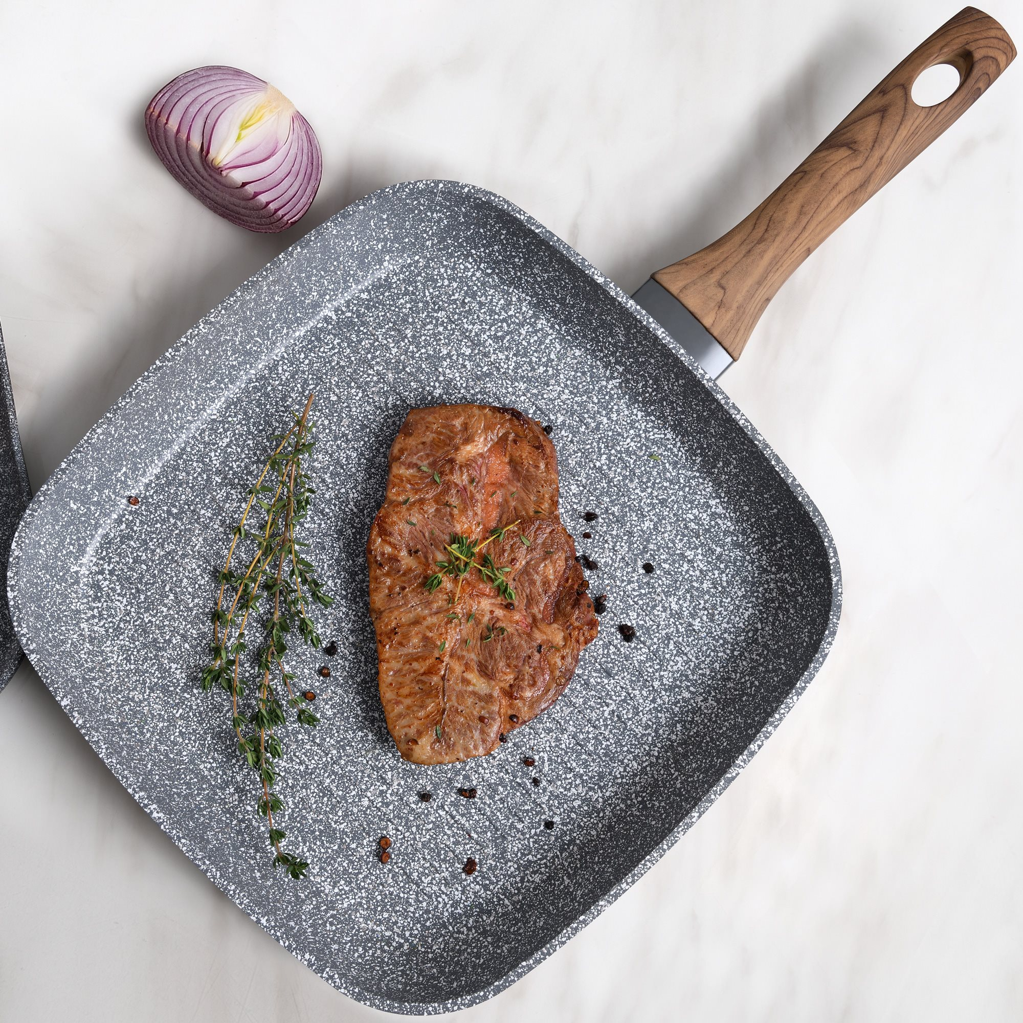 Un bistec cocinado con tomillo y cebolla roja en una sartén cuadrada sobre mármol blanco.