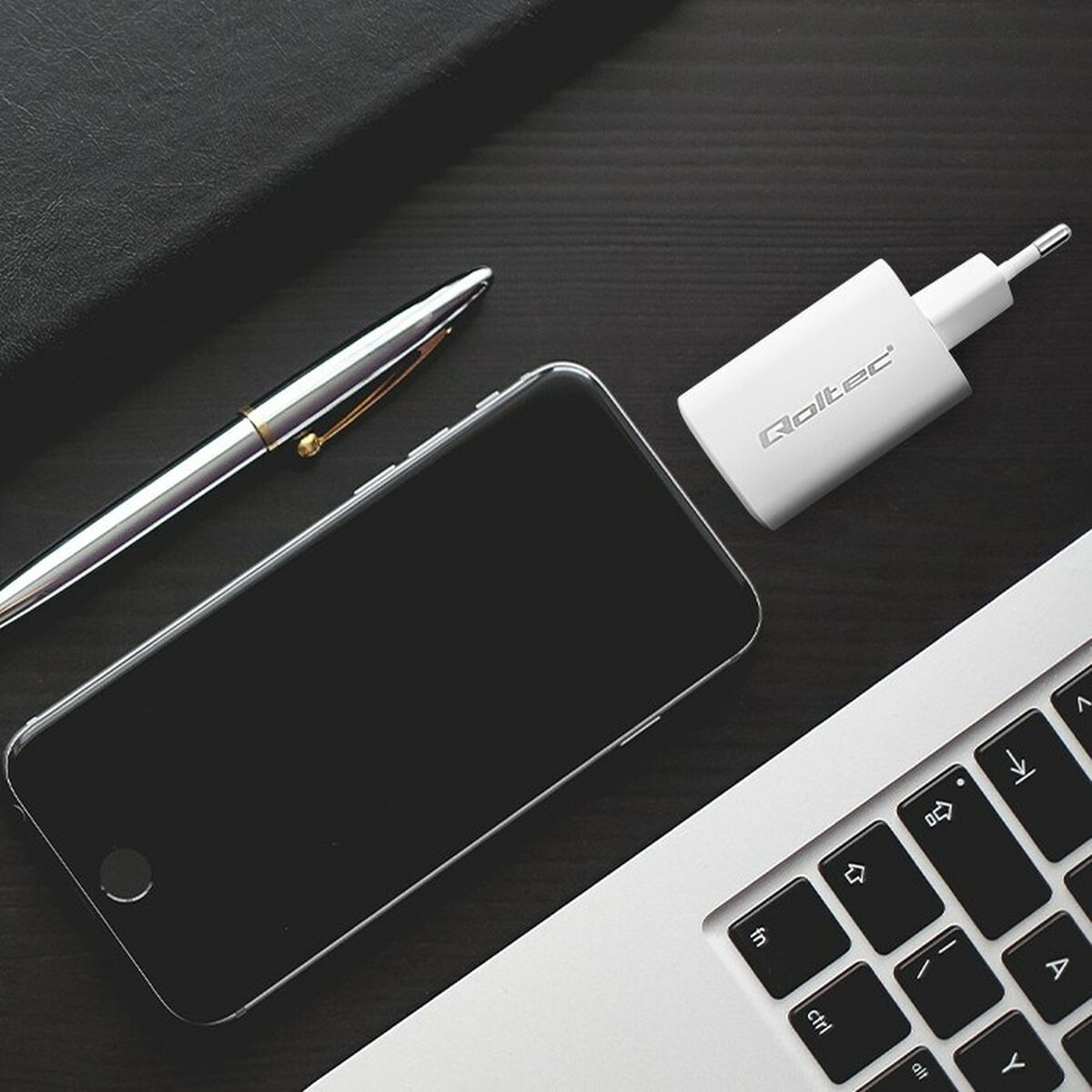 Biała ładowarka, smartfon, długopis i klawiatura laptopa na ciemnym drewnianym biurku.