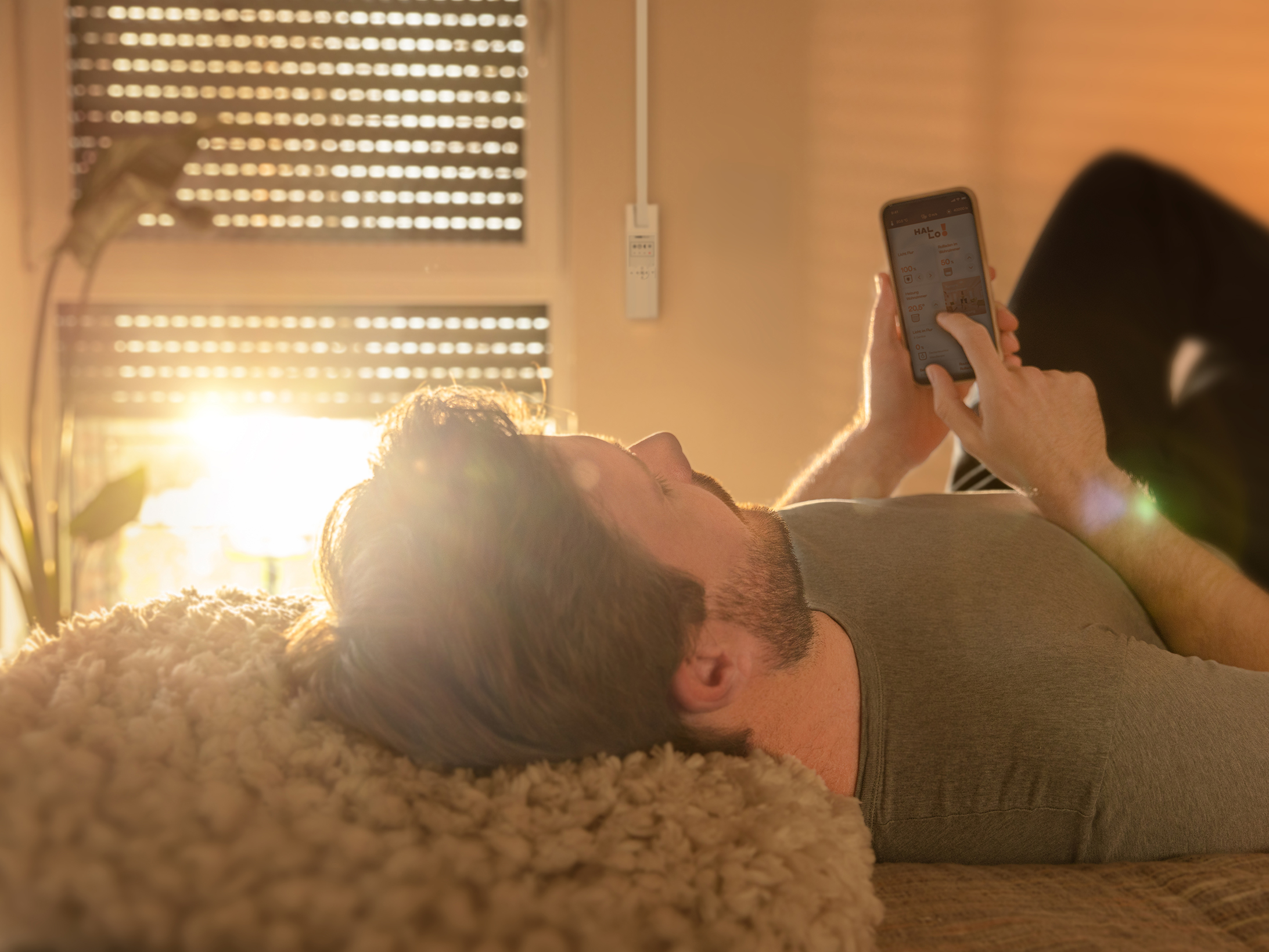 Mann liegt im Bett und schaut auf das Handy, mit Sonnenlicht an der Wand und einem Smart-Home-Hub an der Wand.