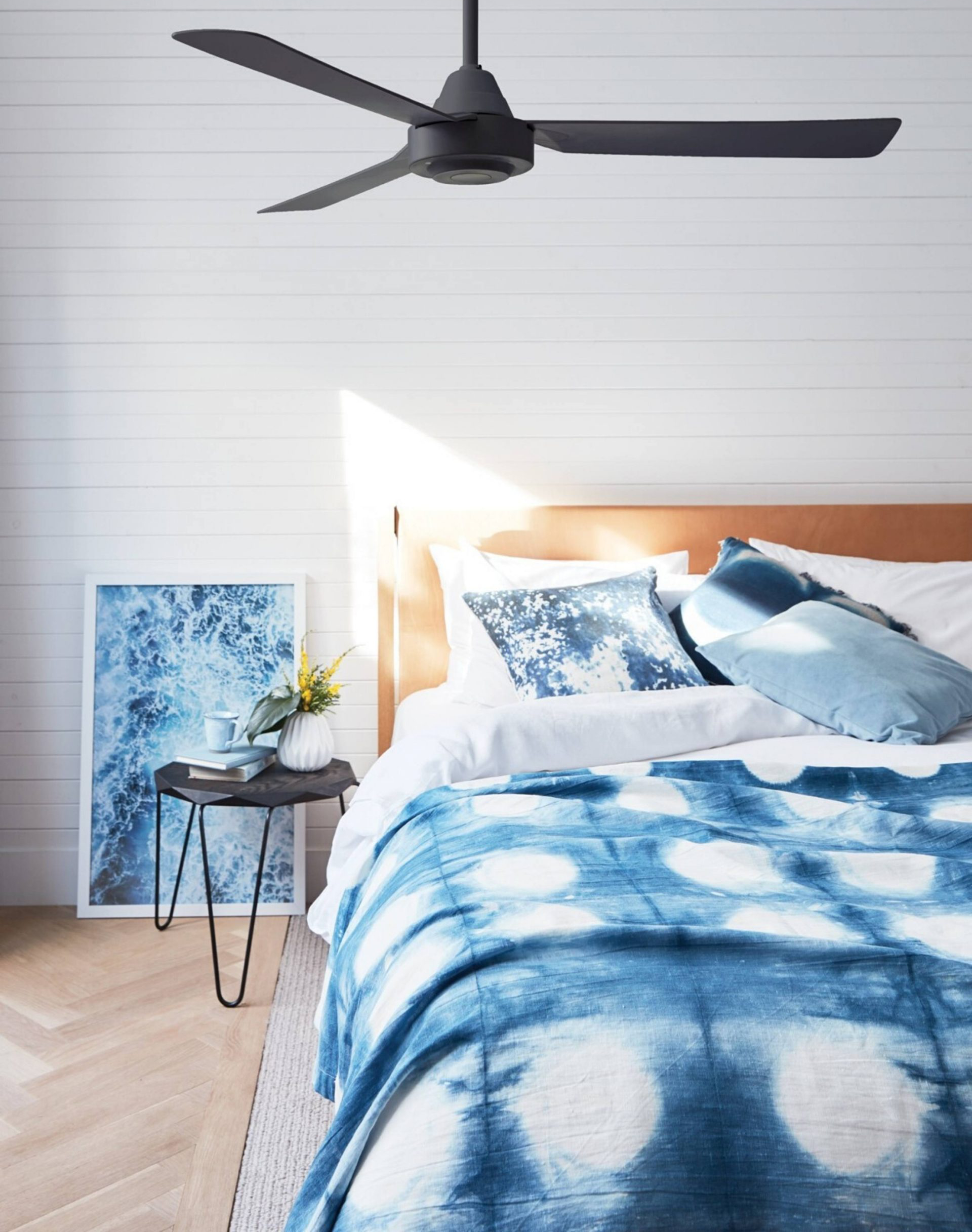 Schlafzimmer mit weißen Wänden, Bett mit blauer Decke und Kissen, ein schwarzer Ventilator und ein Bild.