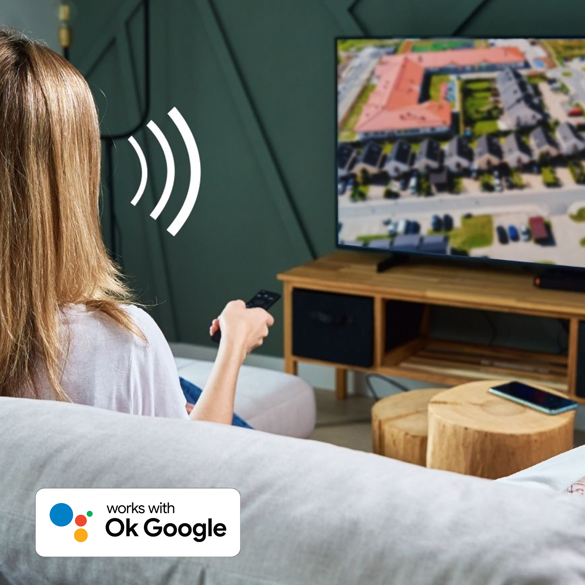 Eine Frau hält eine Fernbedienung. Ein Fernseher ist im Hintergrund. Der Text lautet "works with Ok Google".
