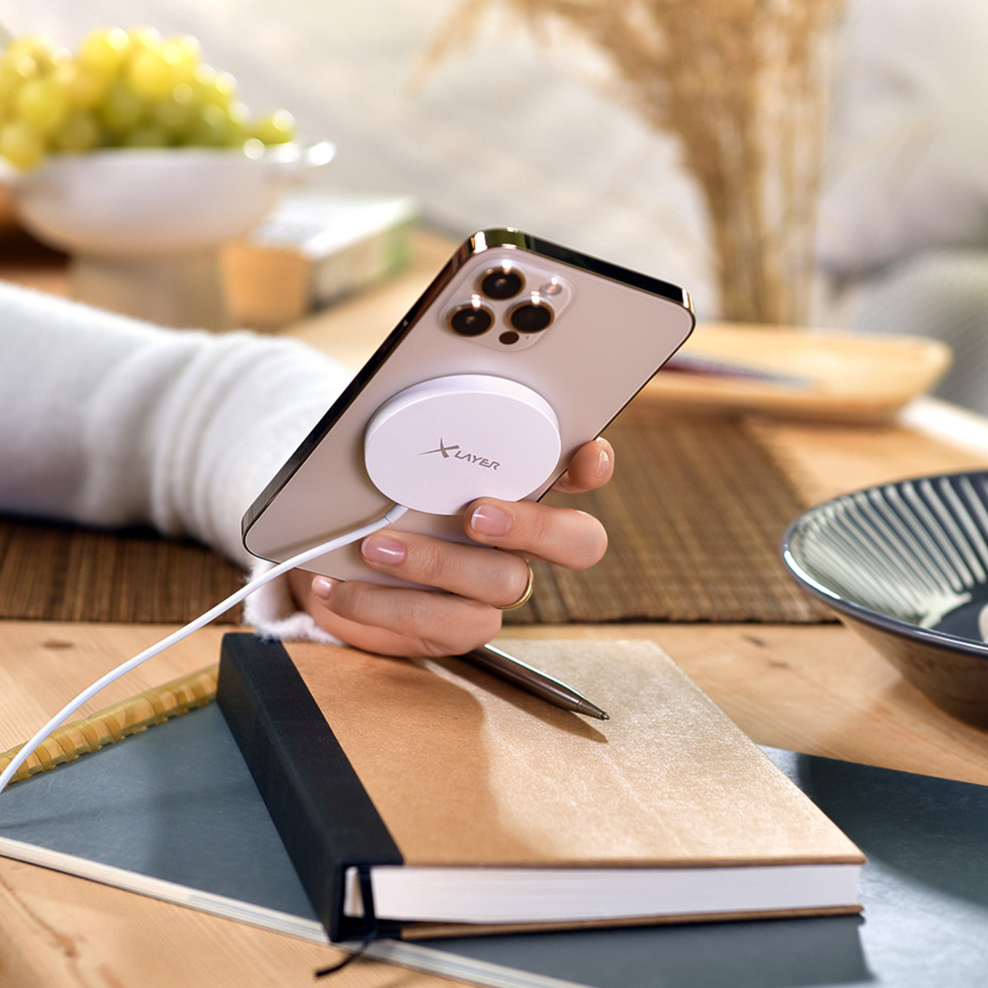 Hand, die ein Telefon mit einem weißen, kreisförmigen Ladegerät hält, auf einem Tisch mit Buch und Stift.