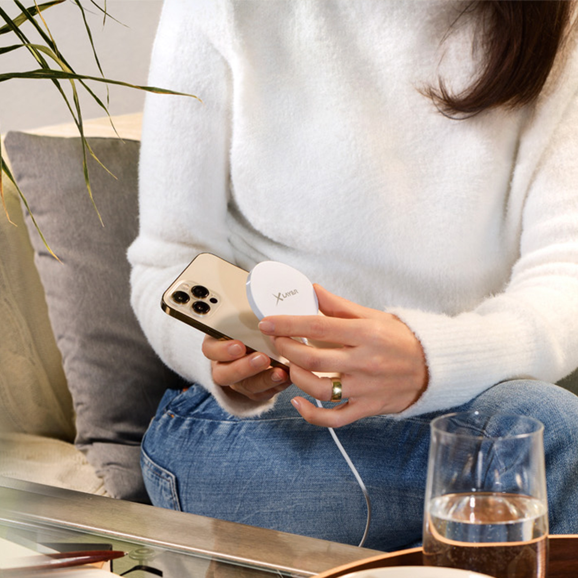 Frau hält Telefon und Ladegerät. Weißer Pullover, Jeans und ein Glas Wasser sind ebenfalls sichtbar.