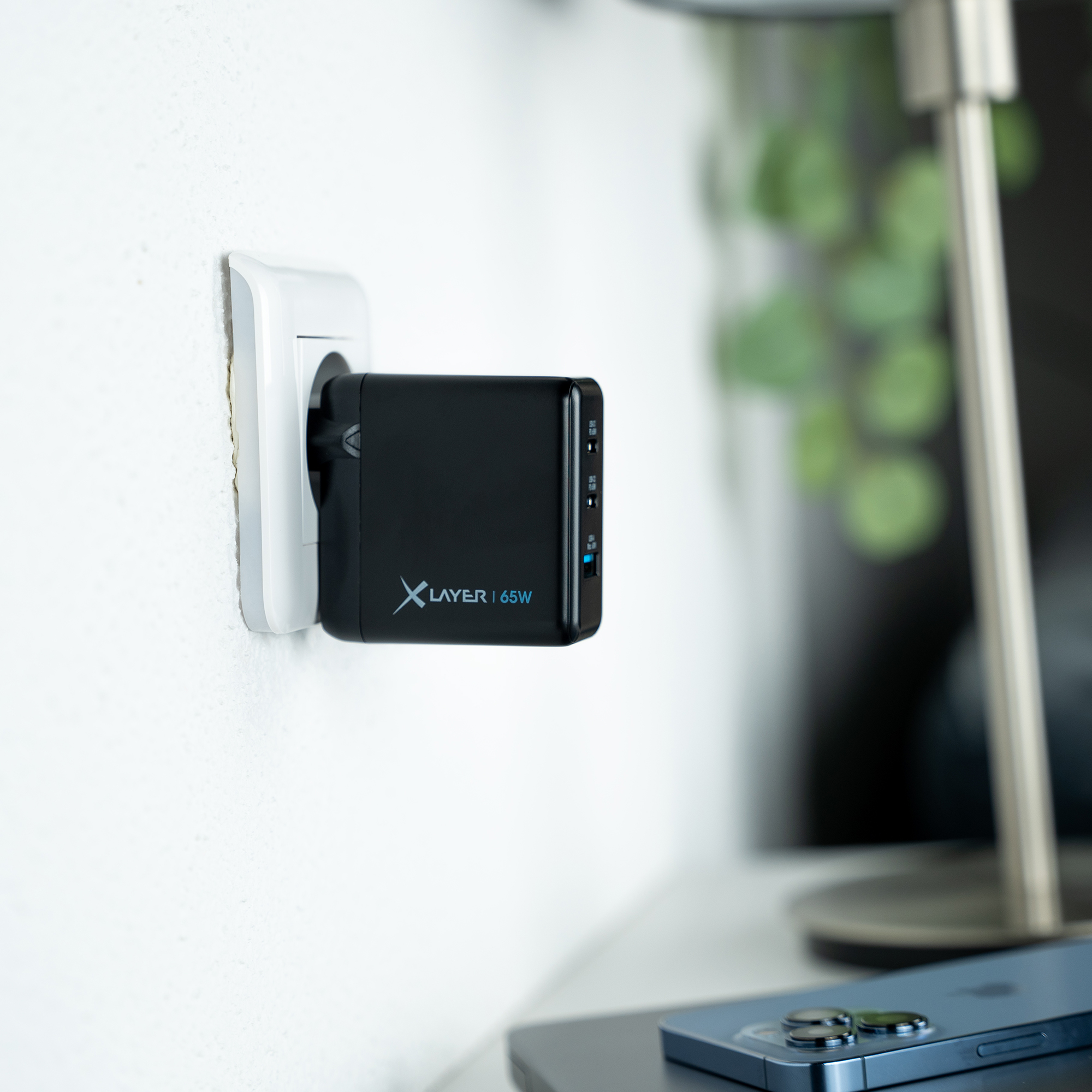 Schwarzer Netzadapter, der in eine Steckdose gesteckt ist, mit einem Telefon daneben. Der Hintergrund ist unscharf.