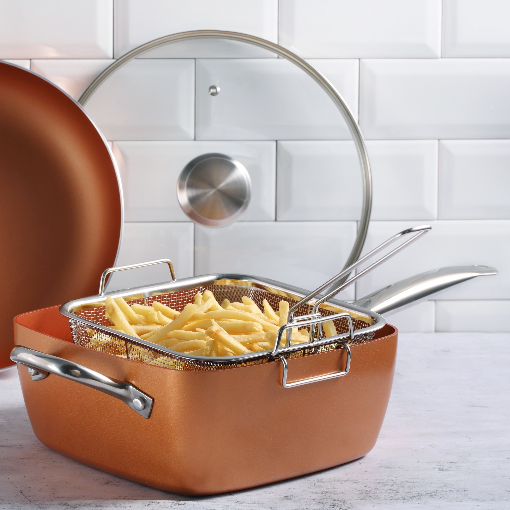 Una freidora de color cobre con papas fritas dentro, una tapa de vidrio y un fondo de azulejos blancos.