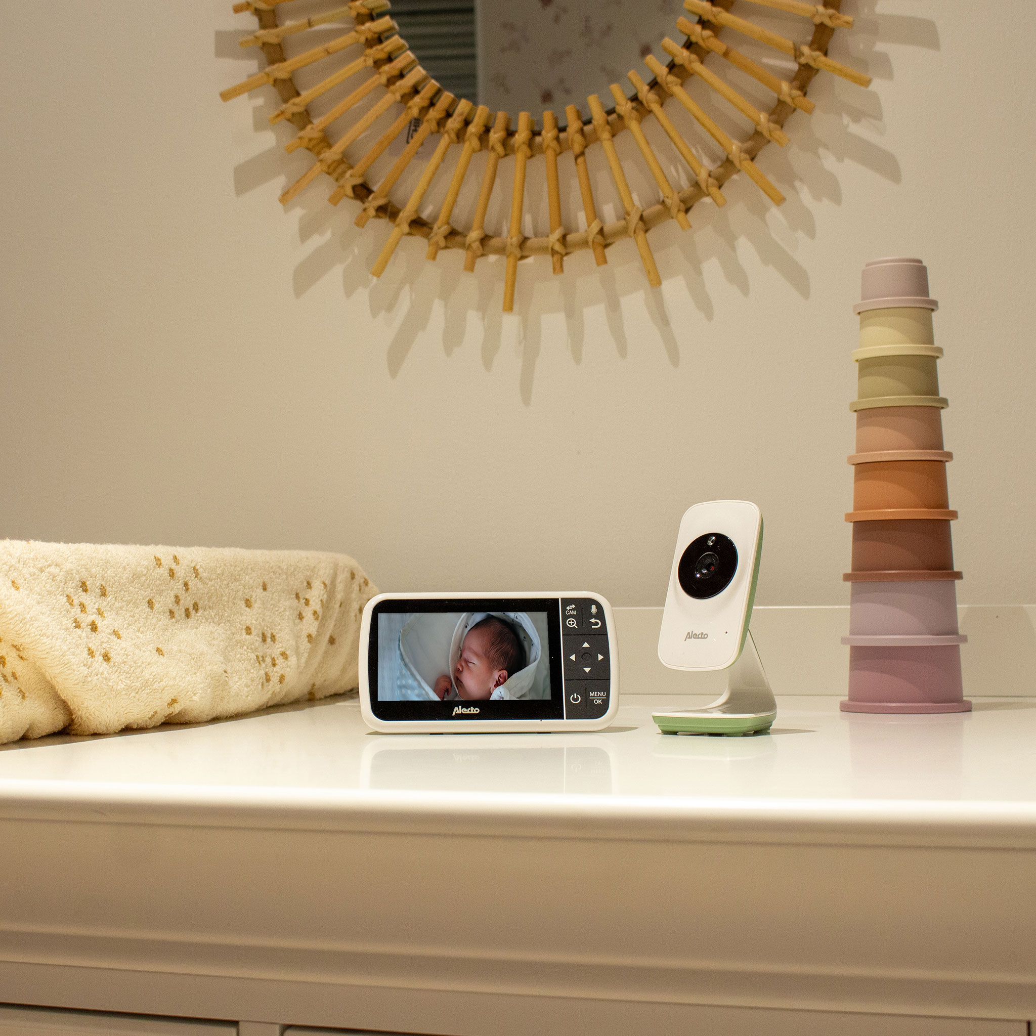 Baby monitor on a white surface with a baby's face on the screen, with a stack of colored cups.