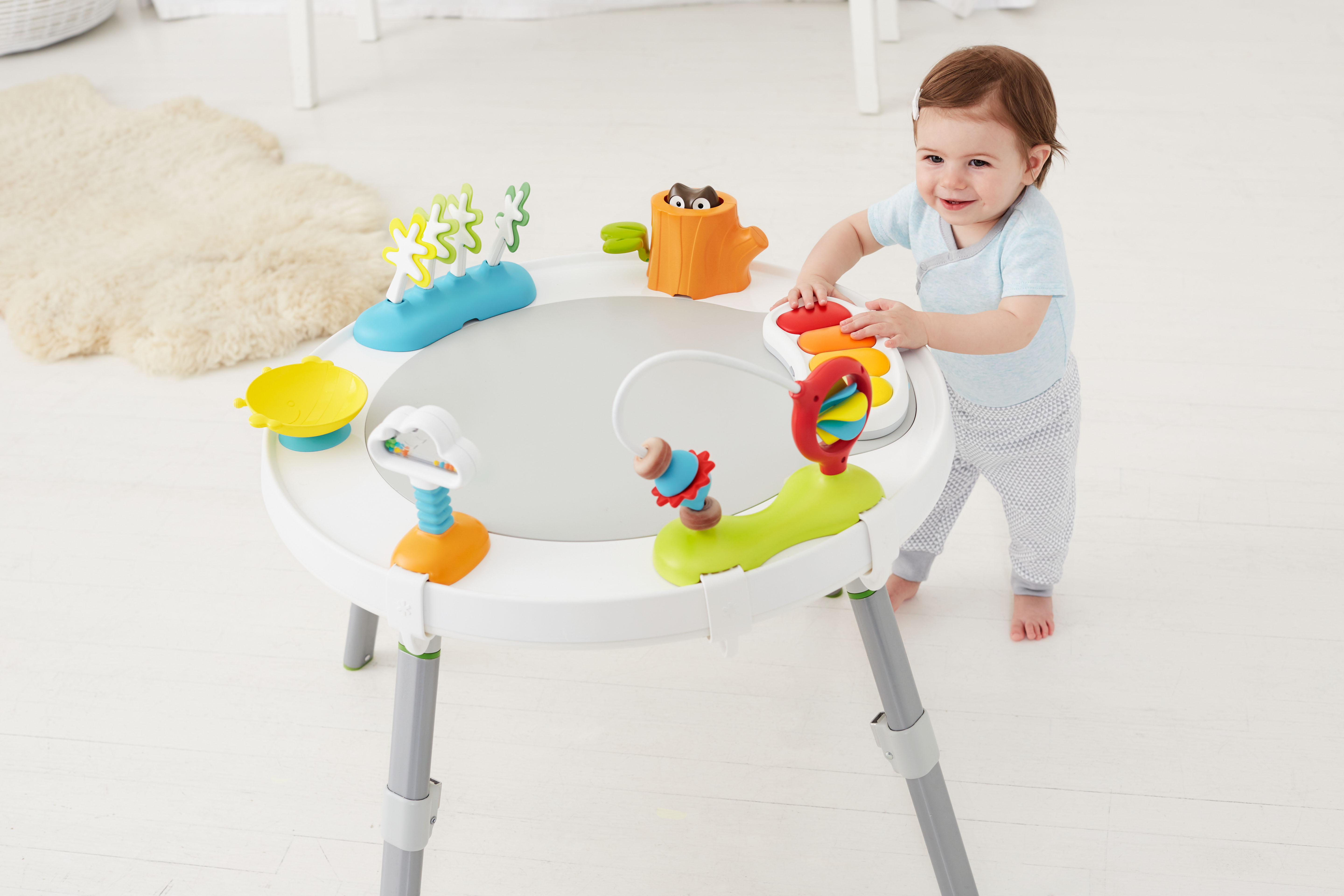 Ein Baby steht an einem Spieltisch mit verschiedenen Spielzeugen. Der Tisch hat eine graue Mitte und steht auf einem weißen Boden.
