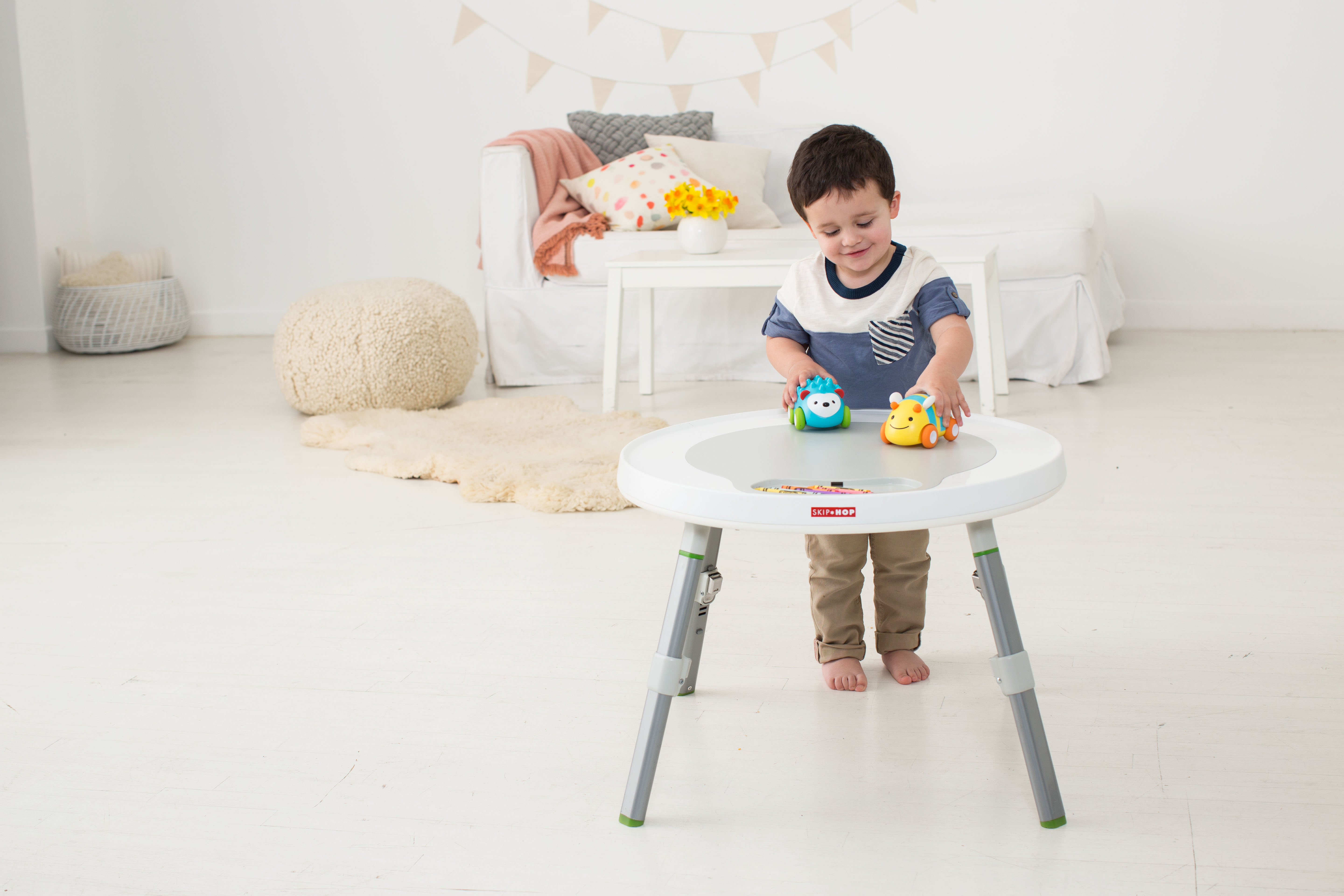 Ein kleiner Junge sitzt an einem Tisch und spielt mit Spielzeugautos. Die Szene spielt in einem hellen, gut beleuchteten Raum.