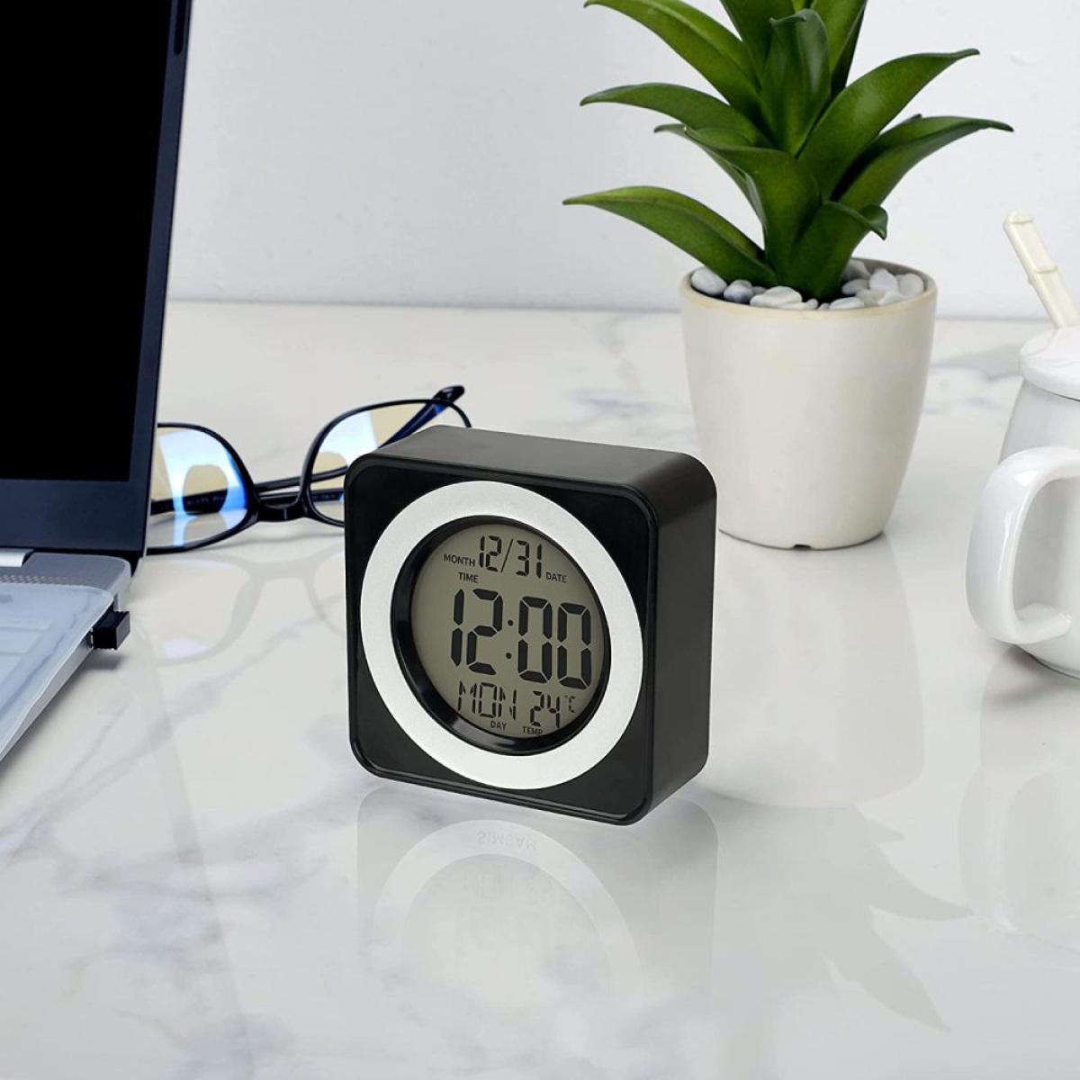 Schwarze Digitaluhr auf weißer Oberfläche. Anzeige zeigt 12:00 Uhr und Datum. Pflanze und Brille sind in der Nähe.