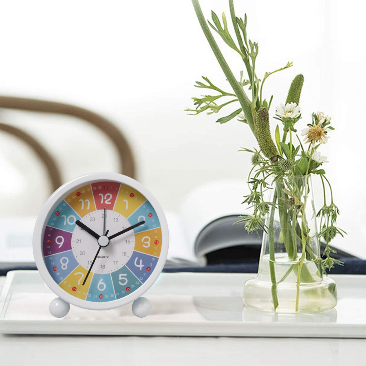 Weißer Wecker mit bunten Zahlen, Blumen in einer Vase auf einem Tablett.