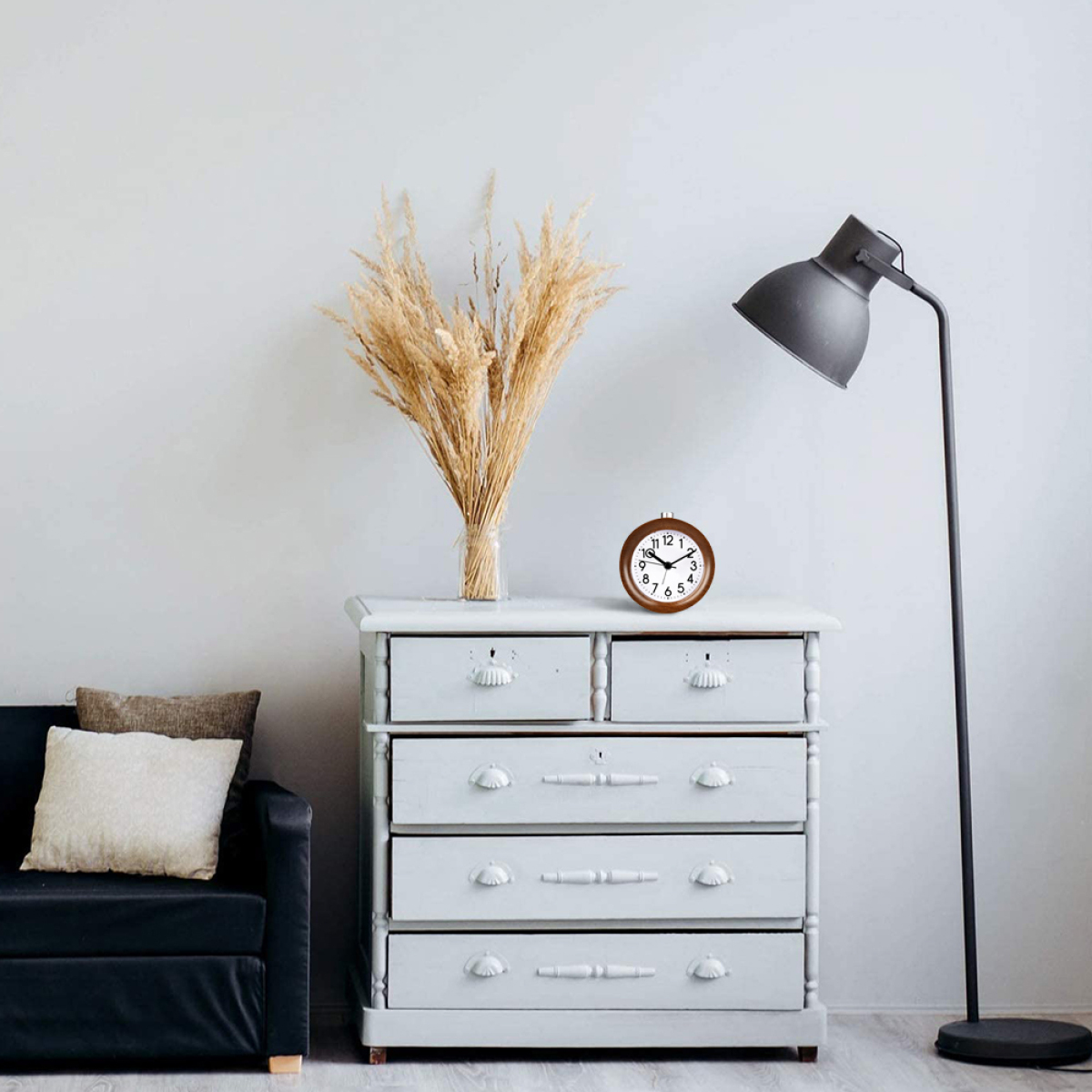 Eine Kommode, ein Blumenstrauß, eine Uhr und eine Lampe vor einer weißen Wand.