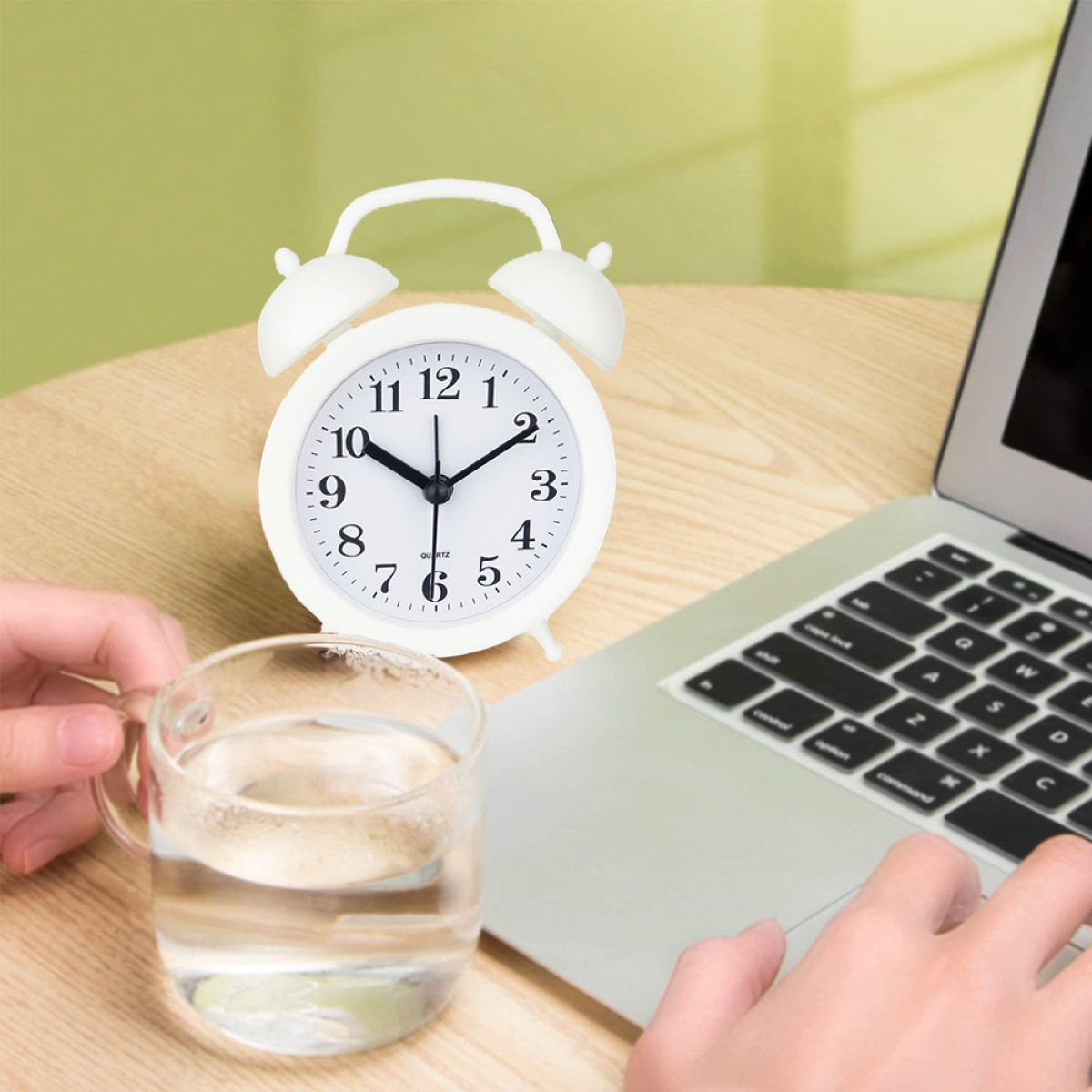Wecker auf Tisch neben Laptop und Glas Wasser, Hand greift nach Uhr.