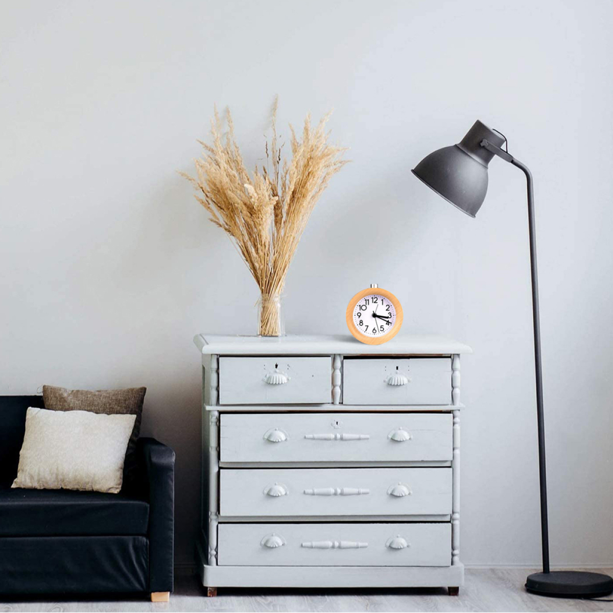 Eine Holzuhr steht auf einer Kommode. Eine Vase mit trockenen Pflanzen, eine Lampe und eine Couch sind zu sehen.