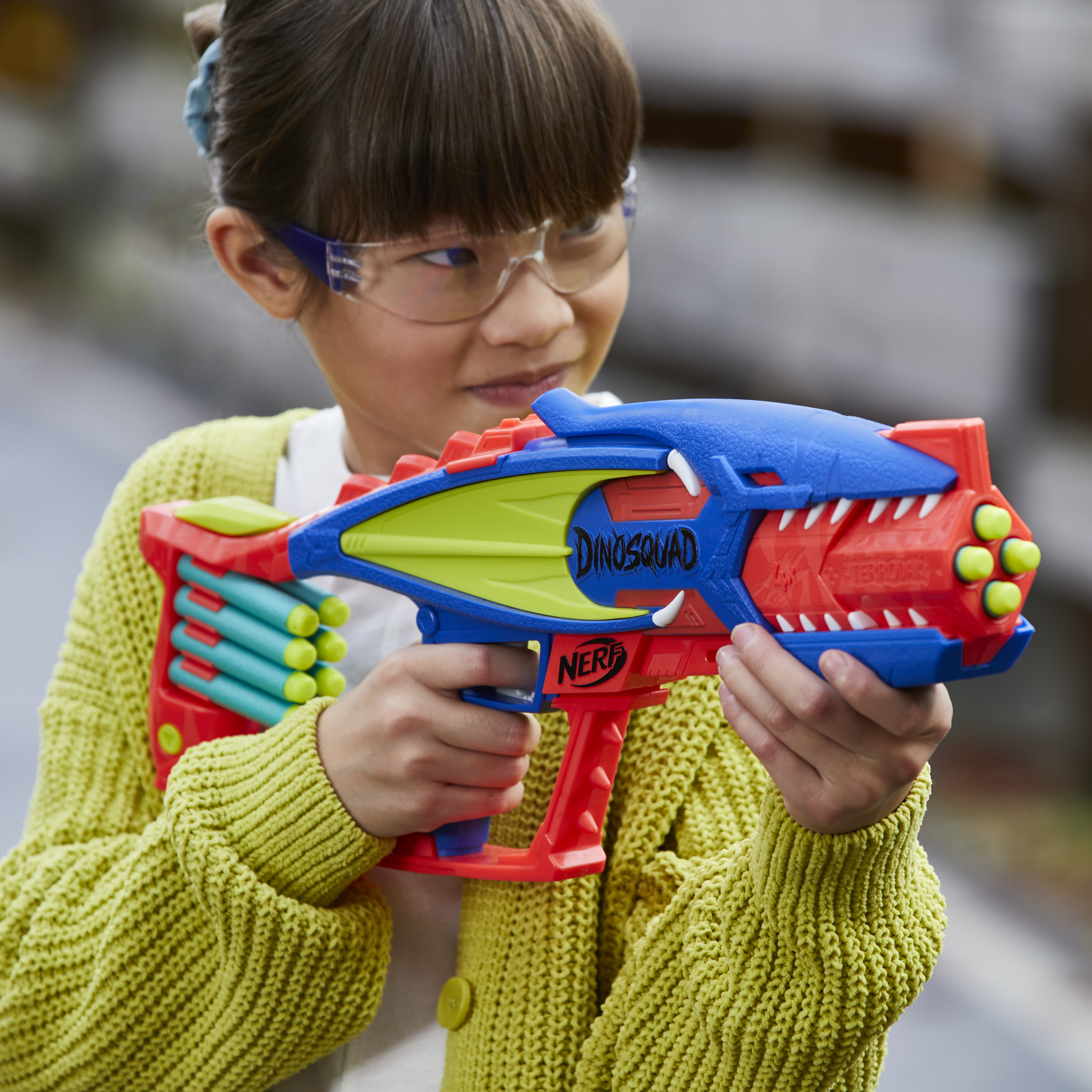 Dziewczyna trzymająca zabawkę w kształcie pistoletu dinozaura, w okularach ochronnych i zielonym swetrze.