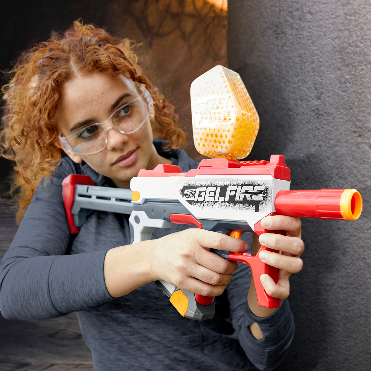 Eine Frau mit Schutzbrille hält eine rot-weiße GelFire-Pistole, mit Gelkugeln in der Luft.