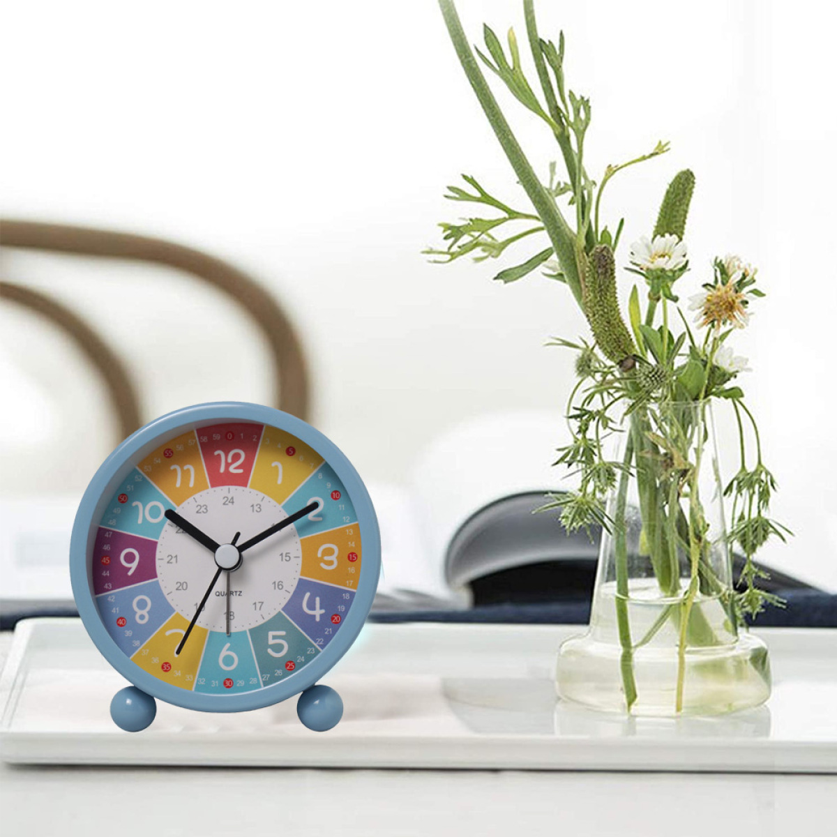 Eine kleine Uhr mit buntem Zifferblatt und eine Glasvase mit Blumen auf einem weißen Tablett.