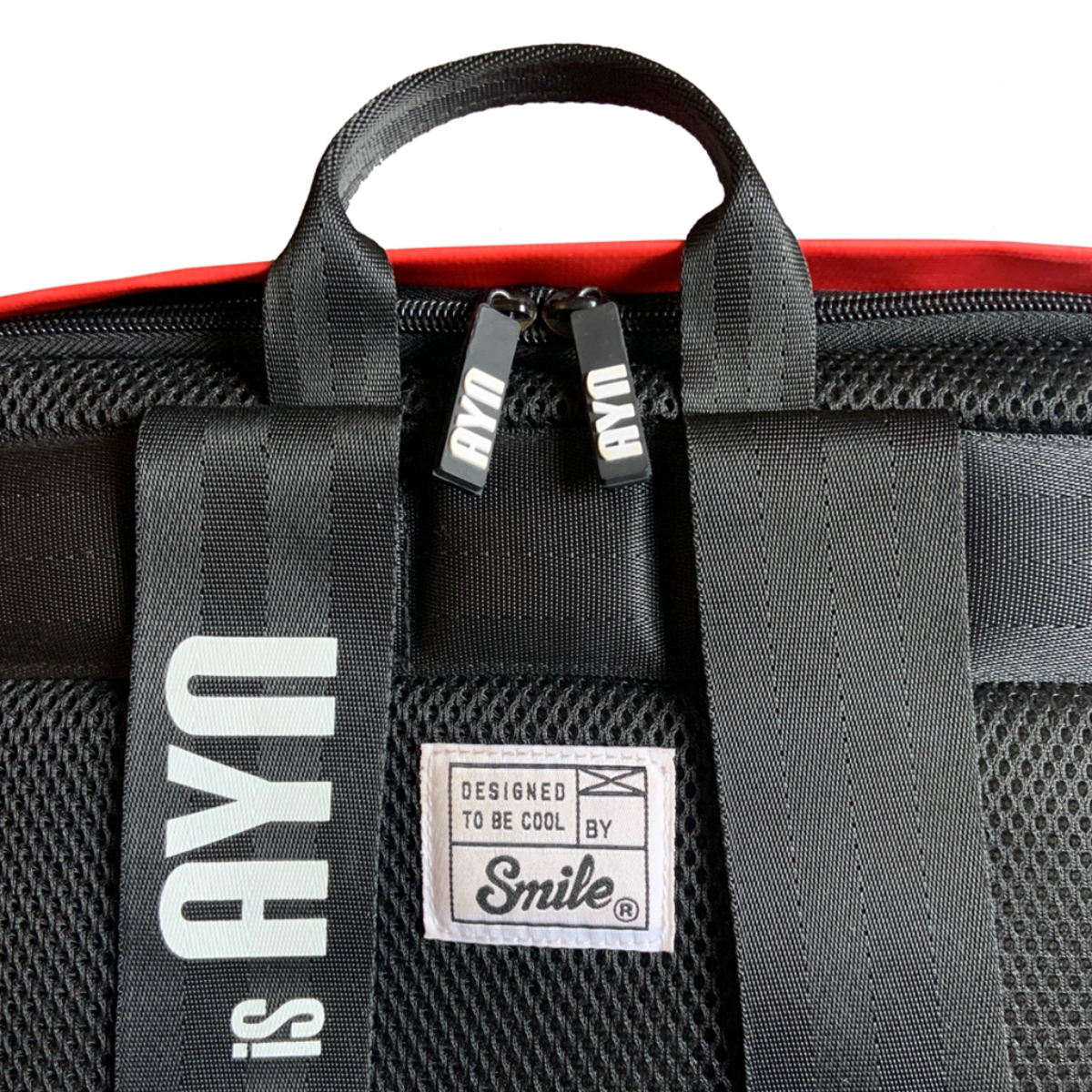 Primer plano de una mochila negra. Correas negras con texto blanco que dice 'AYN'. El logo es 'Designed to be cool by Smile'.