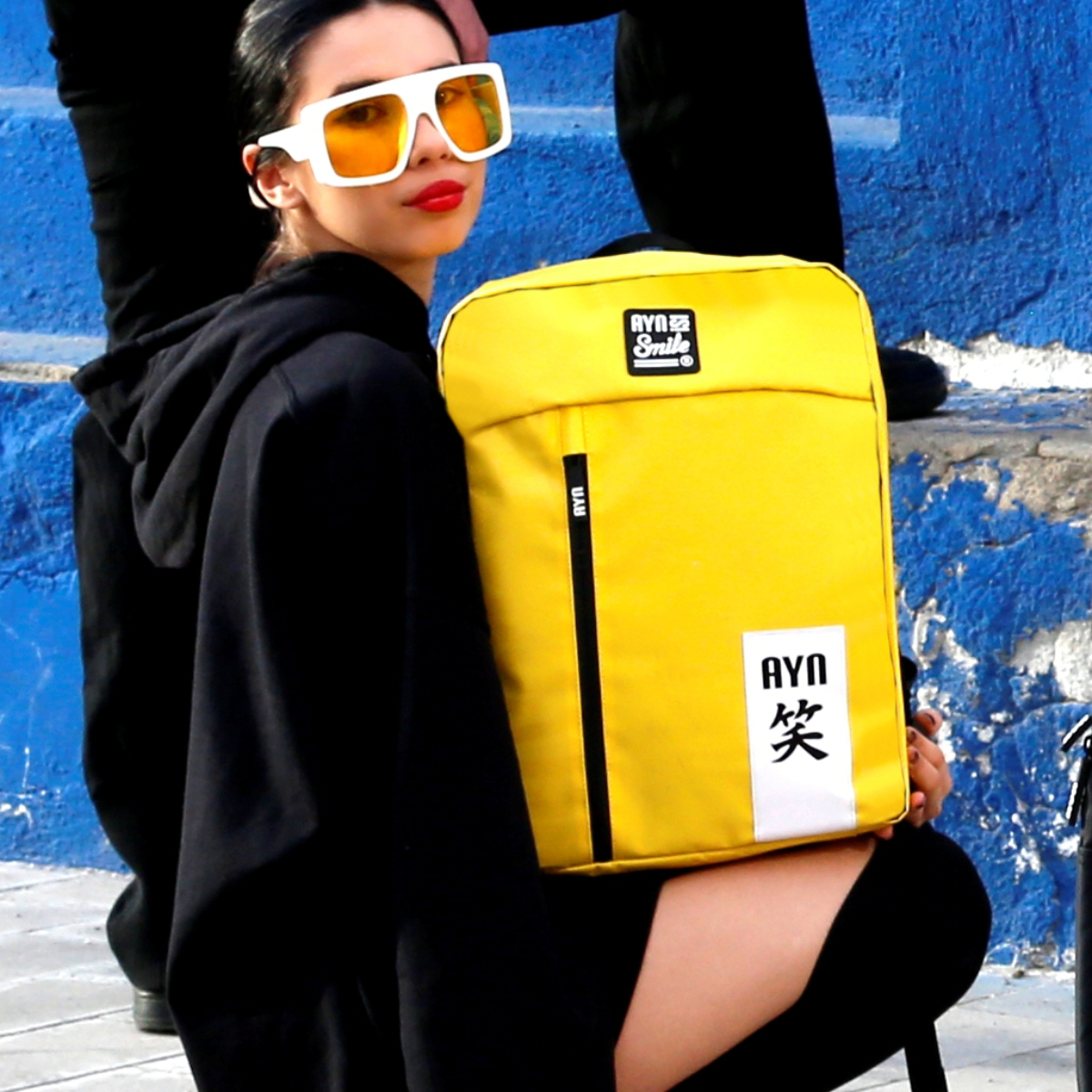 Una mujer con gafas de sol sostiene una mochila amarilla con una raya negra. El fondo es azul.