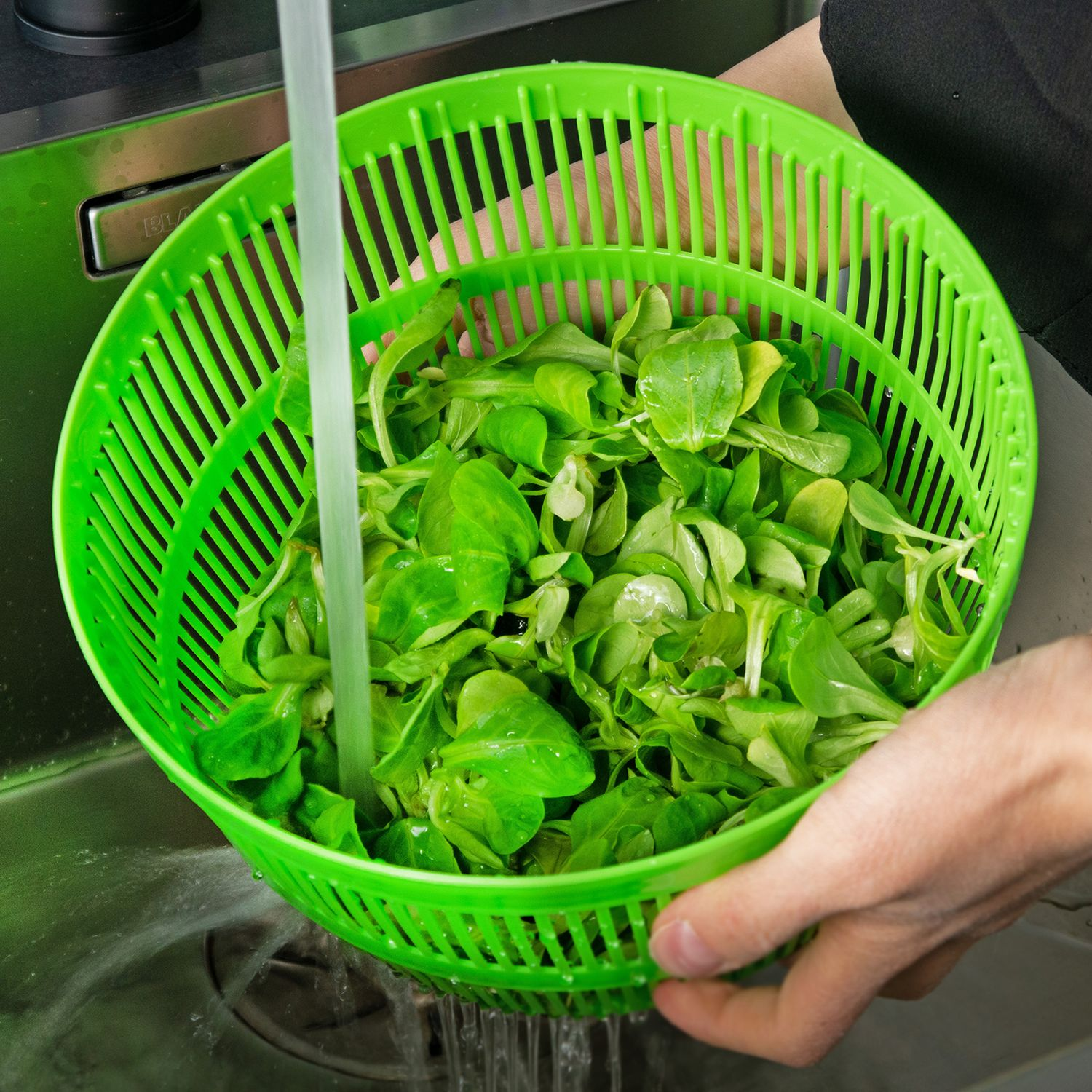 Hände halten ein grünes Sieb mit grünem Salat. Wasser läuft über den Salat.